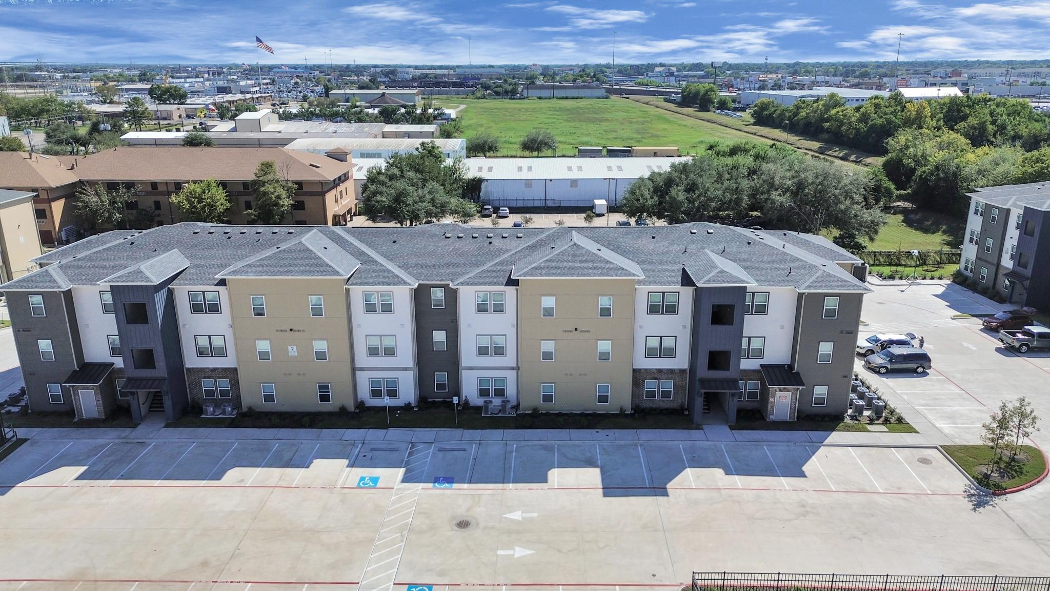 Aerial view of a multi-unit residential building with a mix of brown and beige exterior colors. The building is surrounded by a parking lot with marked spaces. In the background, there are other structures and an expansive green area. Clear blue sky overhead with a few clouds.