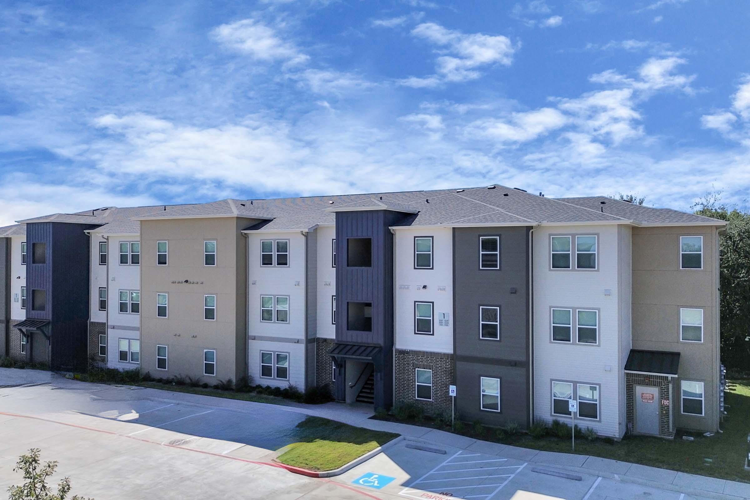 A modern multi-story apartment building with a mix of beige and gray siding. It features three floors with balconies and large windows. The landscape includes a well-maintained lawn and a parking area with a designated accessible parking space. The sky is partly cloudy.