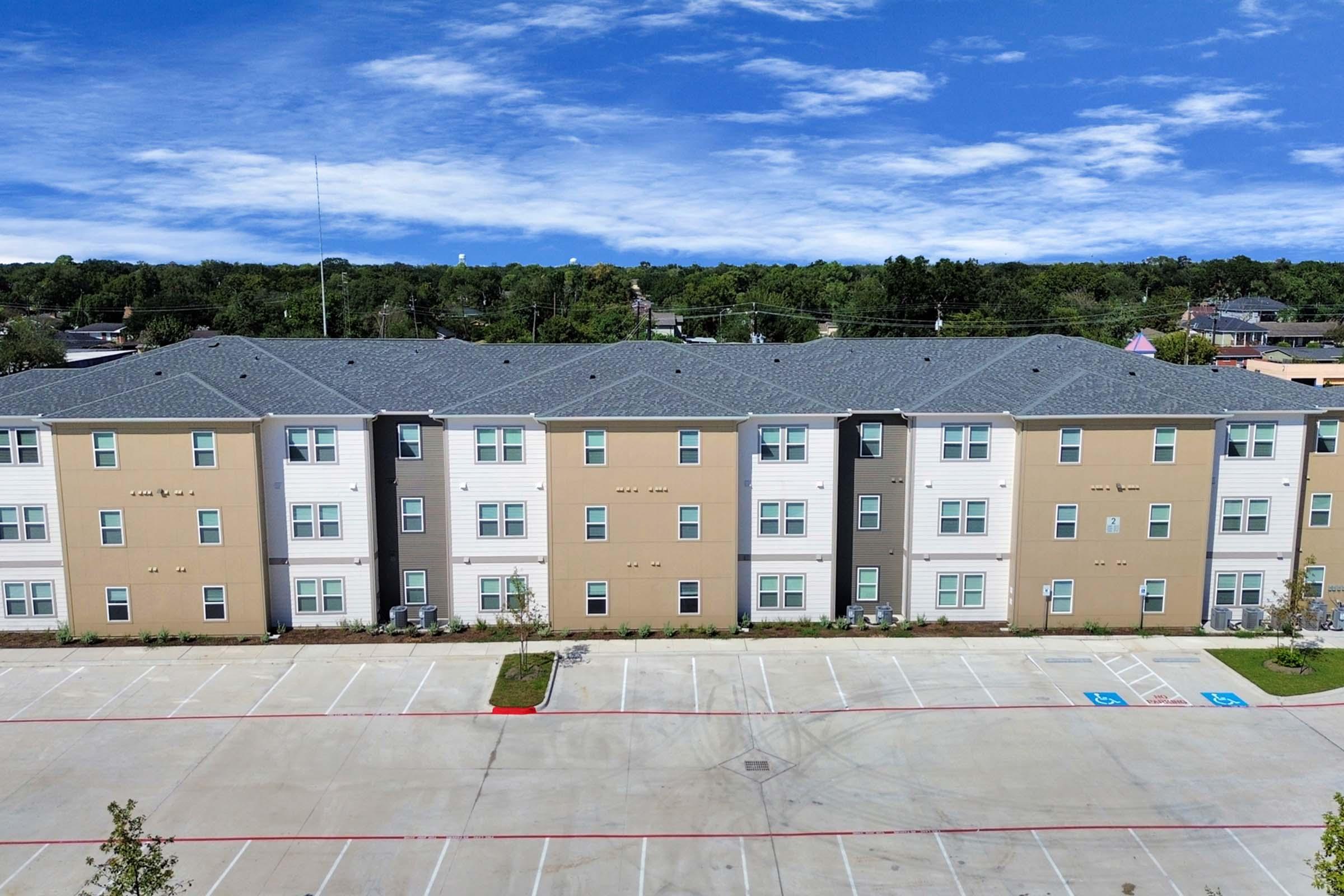 A row of modern apartment buildings featuring a mix of beige and white exteriors, surrounded by green trees. The parking lot in front has designated spaces, and the sky is clear with a few clouds.