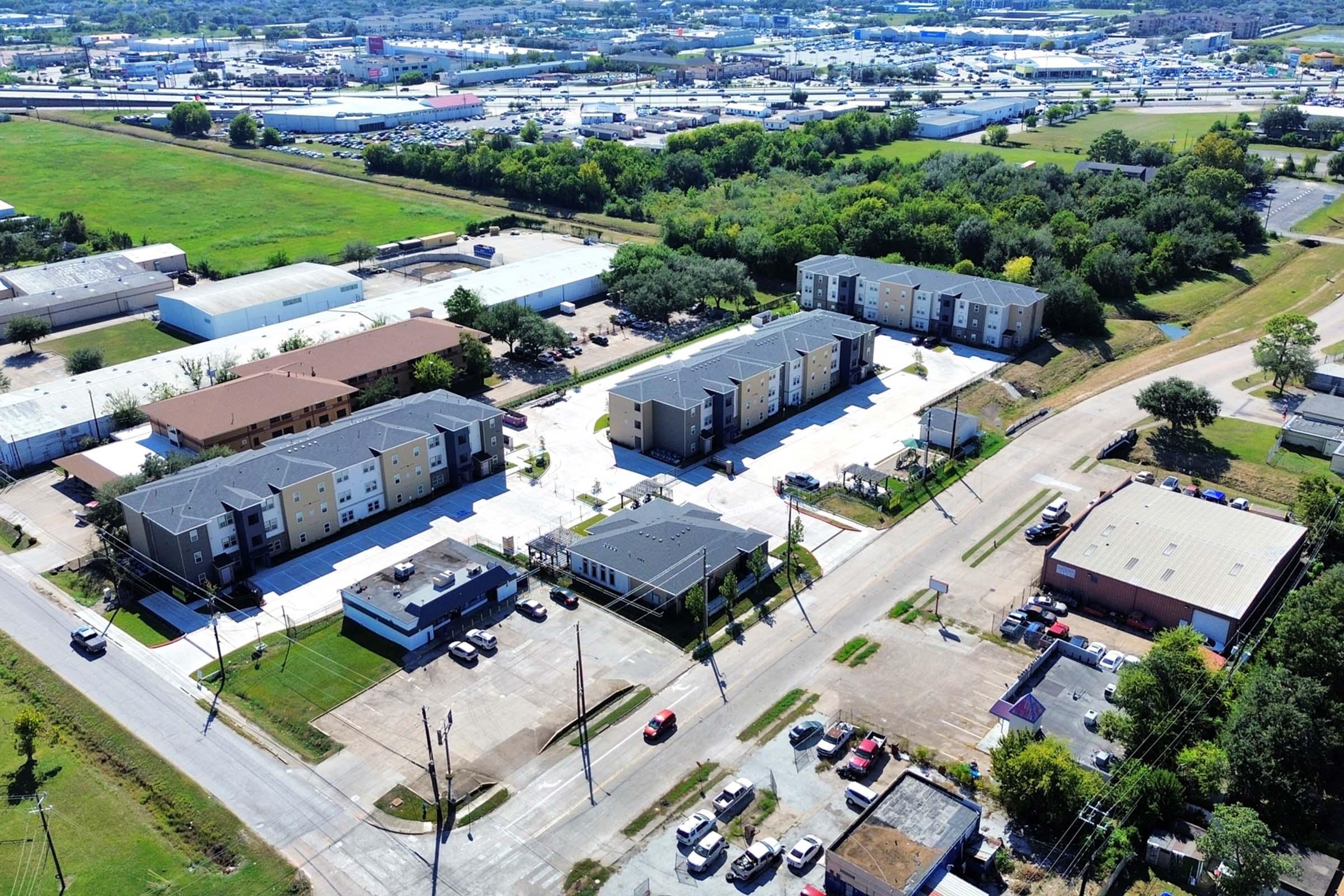 An aerial view of a suburban area featuring multiple residential buildings, parking lots, and nearby commercial spaces. The landscape includes green fields and industrial structures in the background, showcasing a blend of urban and rural environments.