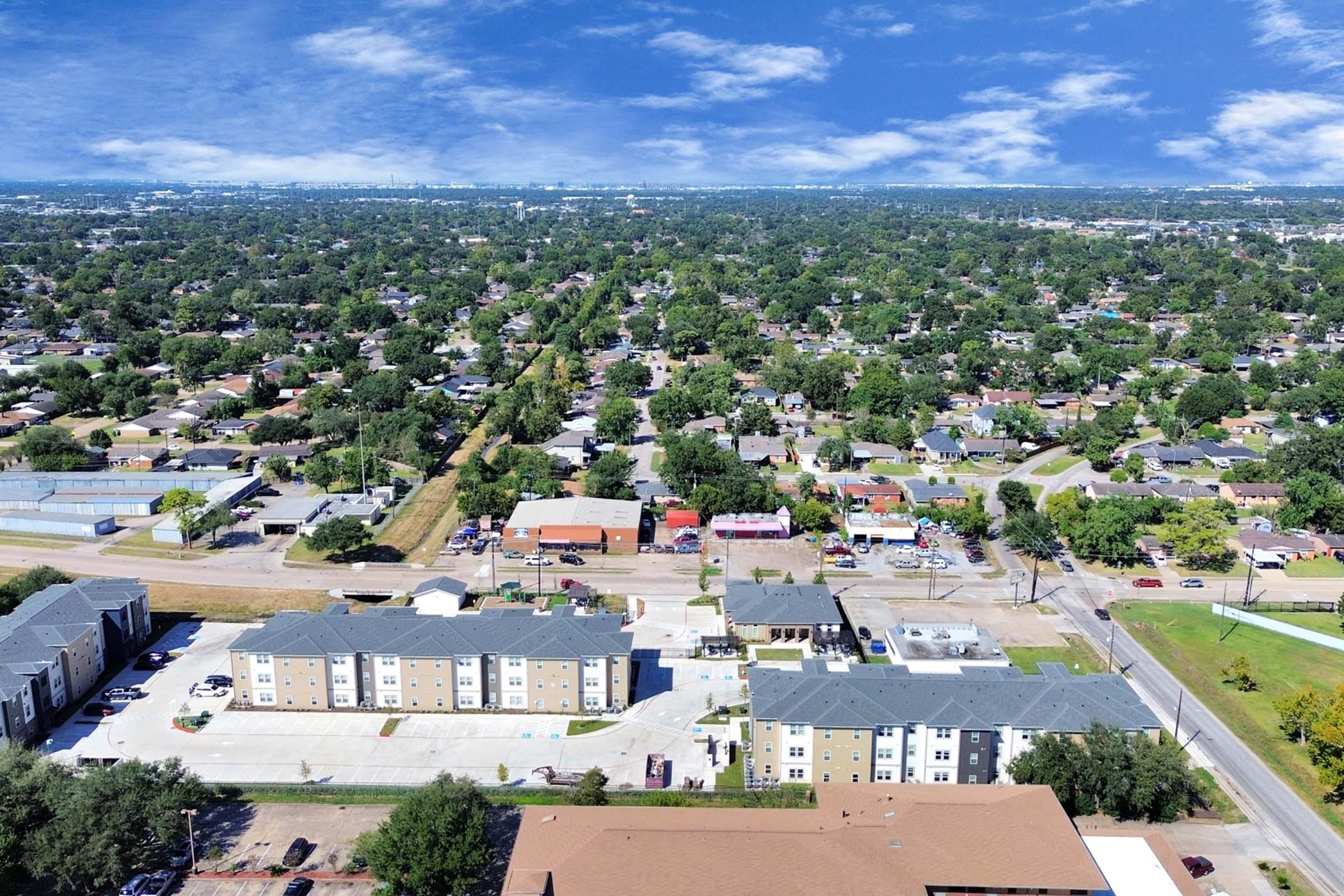 Aerial view of a suburban area featuring a mix of residential homes and commercial buildings. Lush green trees are visible alongside streets, with a few businesses and parking lots in the foreground. The sky is partly cloudy, creating a bright and welcoming atmosphere.