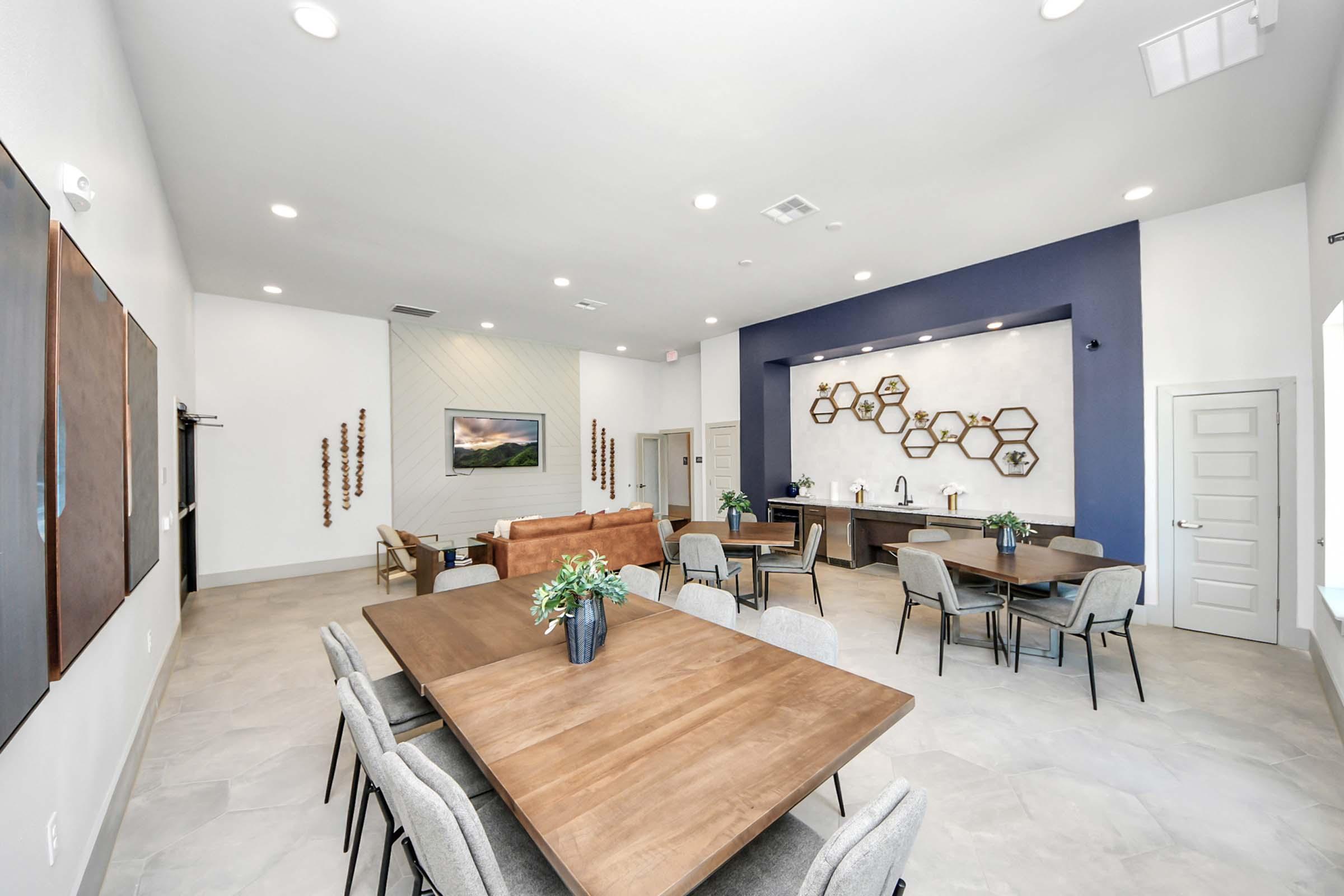 Spacious, modern common area featuring a large wooden dining table with grey chairs, a comfortable couch, decorative wall art, and a coffee station. Brightly lit with natural light, the room has a blue accent wall and stylish decor elements, creating an inviting atmosphere for gatherings or relaxation.