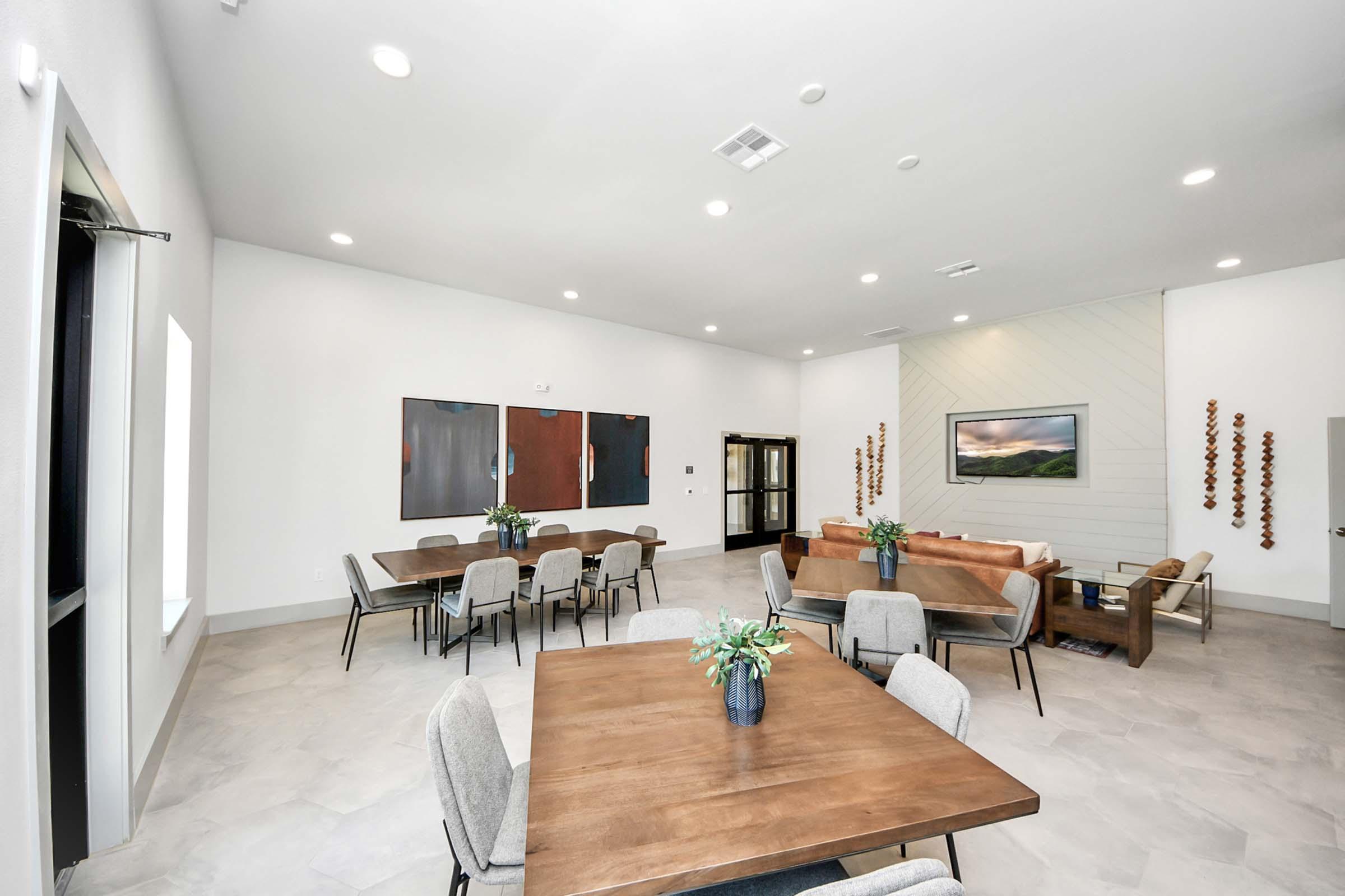 A spacious indoor gathering area featuring modern furniture, including wooden tables with chairs, a cozy couch, and wall art. Natural light floods in through large windows, creating a welcoming atmosphere. The minimalist design includes neutral colors and clean lines, ideal for socializing or meetings.