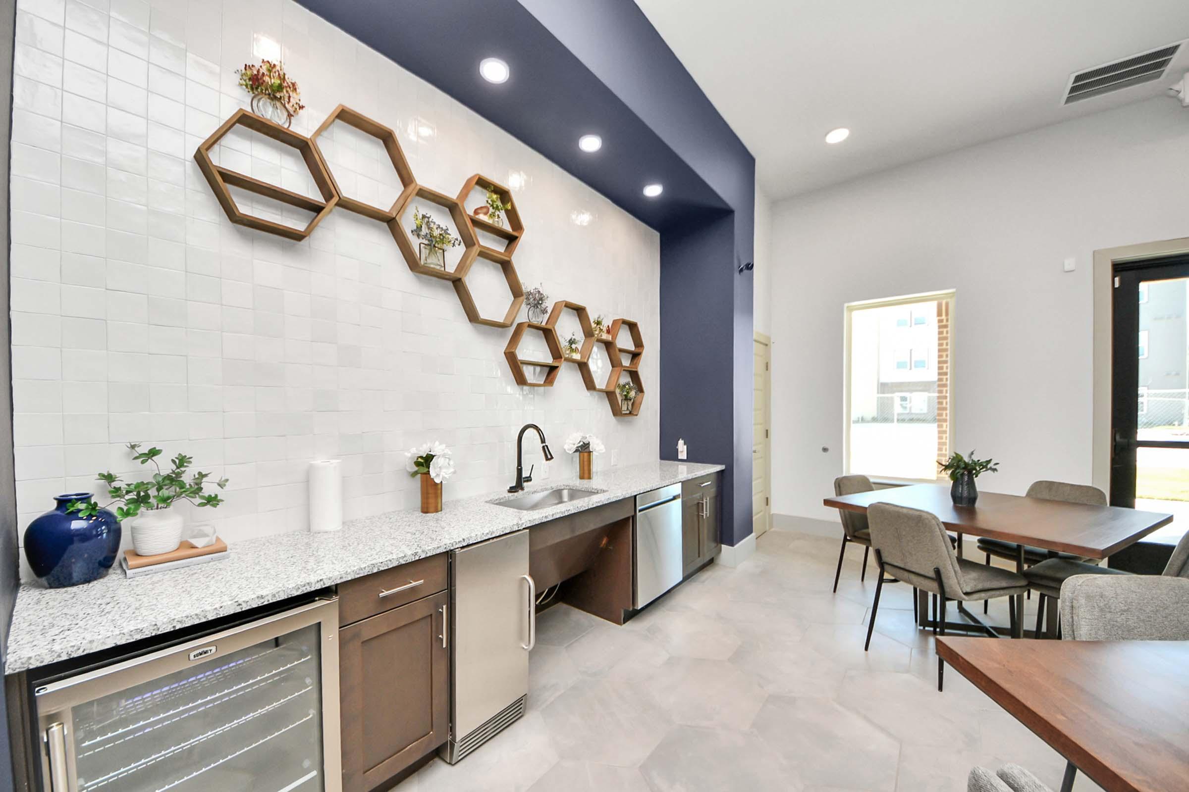 Modern kitchen featuring a marble countertop with a sink and beverage cooler. Honeycomb-shaped shelves on the wall are decorated with plants. Light-colored cabinets complement the design, and there are dining tables and chairs nearby. Natural light streams in from a large window.