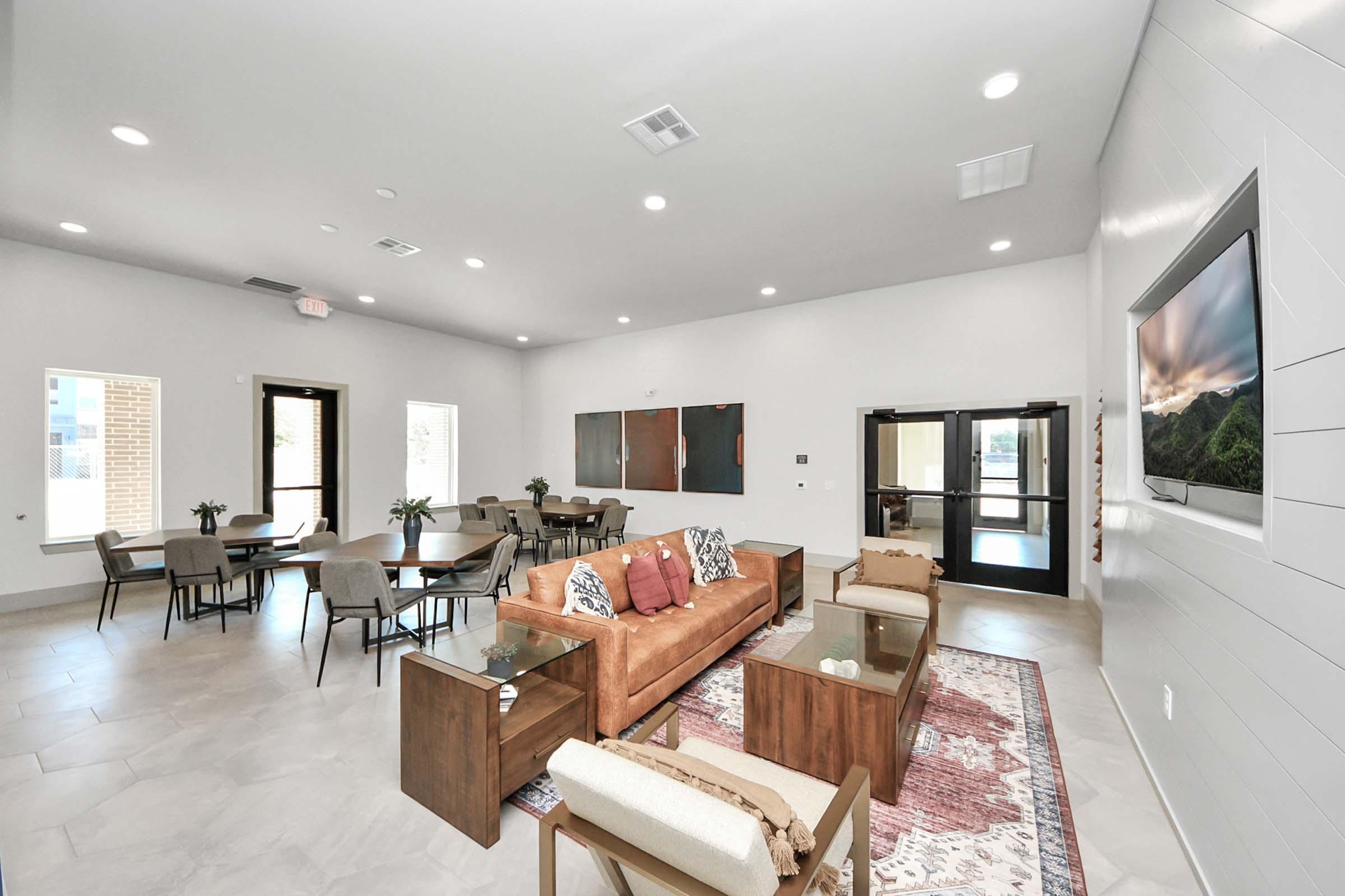 A modern indoor lounge area featuring a cozy seating arrangement with a brown sofa, two armchairs, and a decorative rug. Tables and chairs are arranged for dining or socializing. Large windows allow natural light, and there is a flat-screen TV mounted on the wall, creating a welcoming atmosphere.