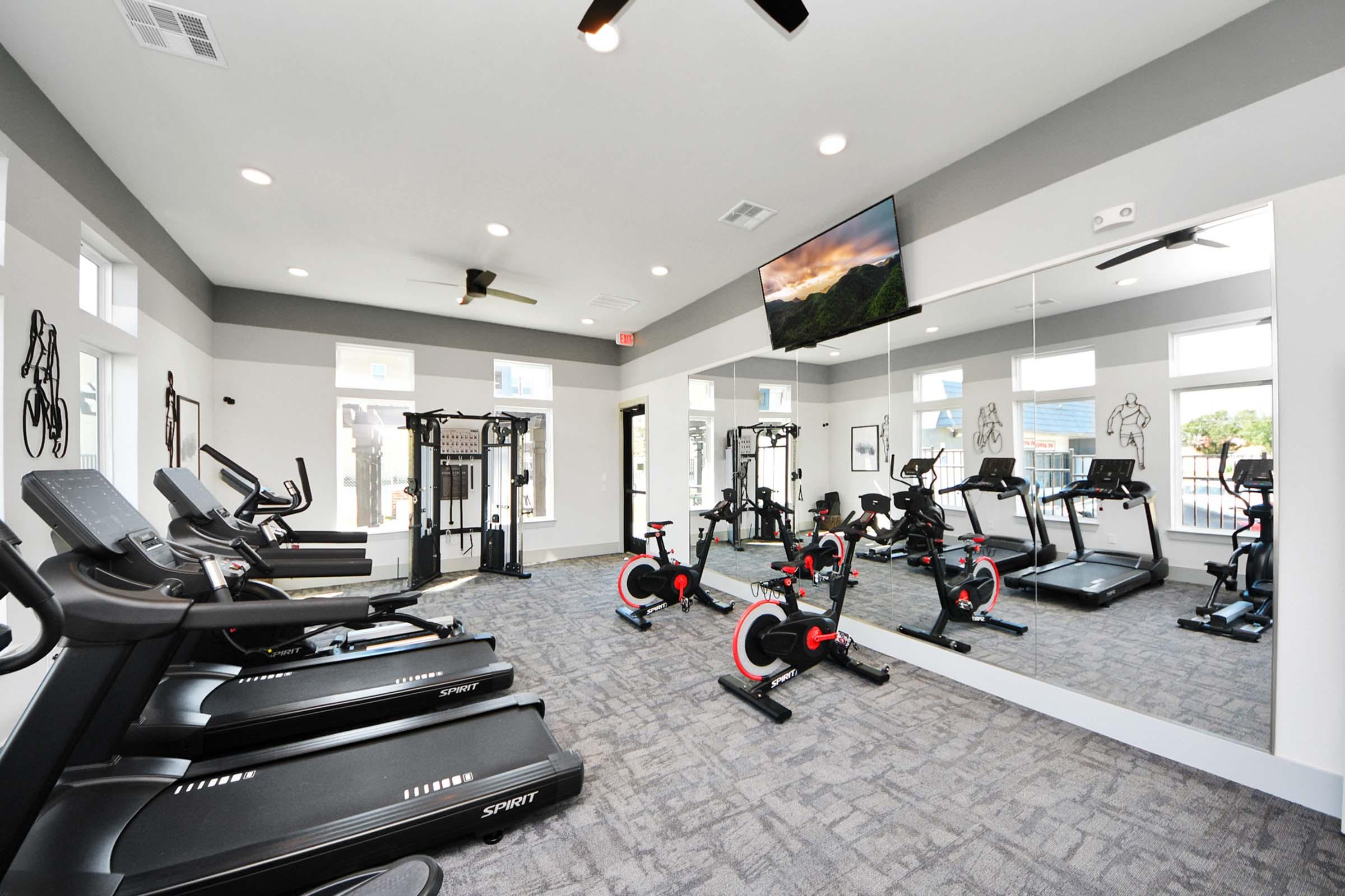 A modern fitness room featuring multiple treadmills and stationary bikes, along with weight training equipment. Large mirrors line the walls, and a flat-screen TV is mounted on the wall. The space is well-lit and has a clean, contemporary design suitable for workouts.