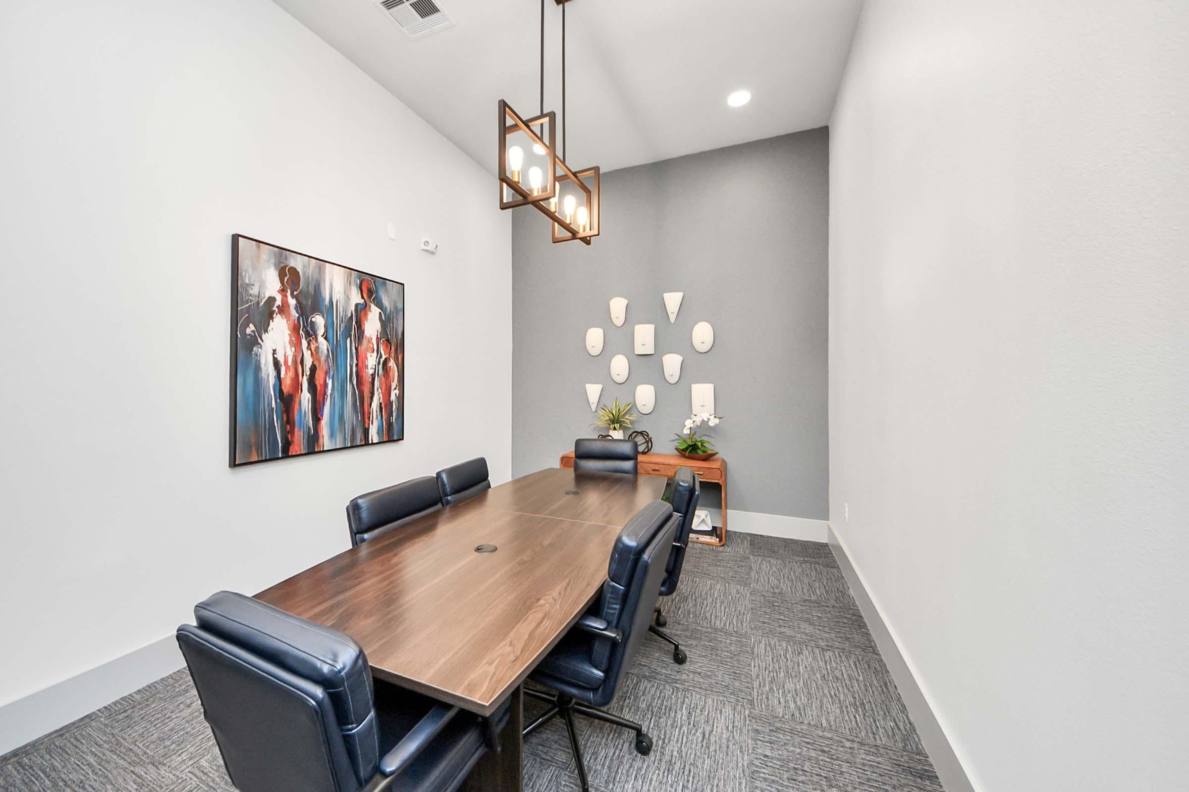 A modern conference room featuring a large wooden table surrounded by dark blue chairs. On the walls, there is a colorful abstract painting and a display of circular wall art. The space is well-lit with a stylish pendant light, and there are plants adding a touch of greenery.
