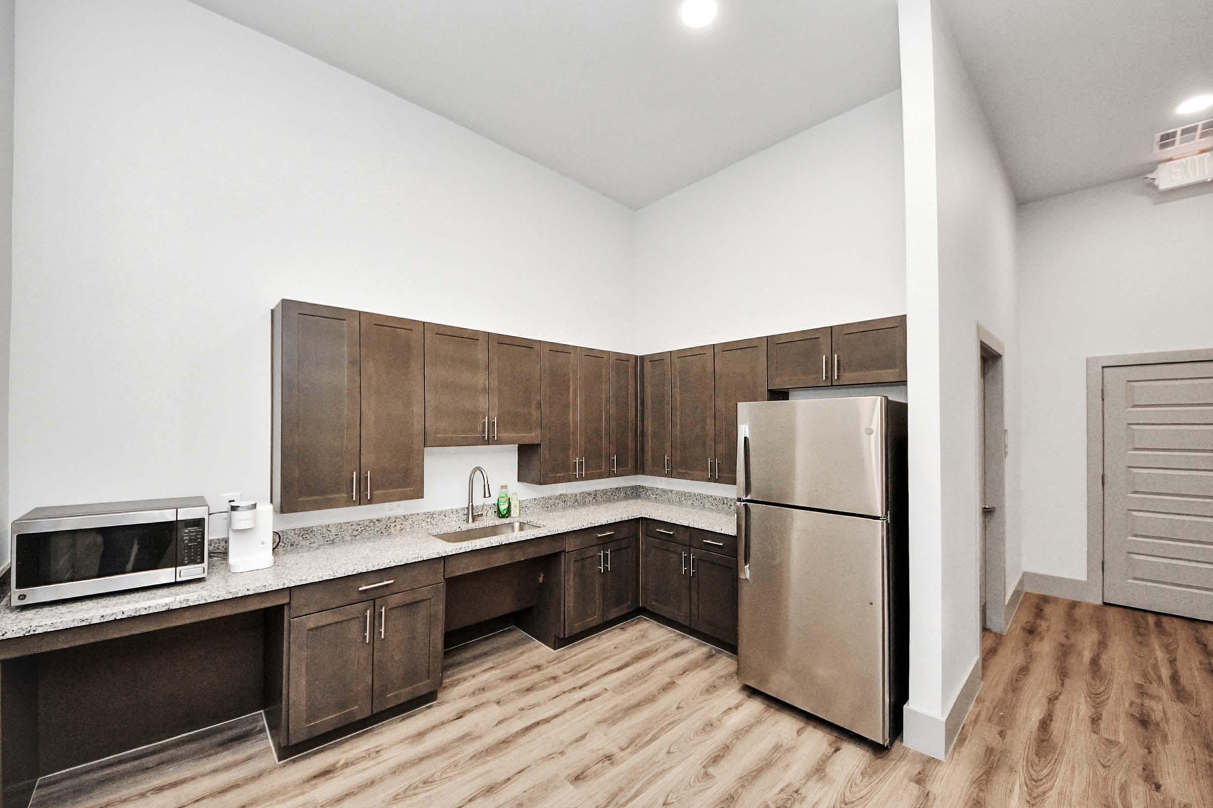 Modern kitchen featuring dark wood cabinets, a stainless steel refrigerator, a microwave, and granite countertops. The space has a light, airy feel with high ceilings and warm wood flooring, creating a contemporary and inviting atmosphere.
