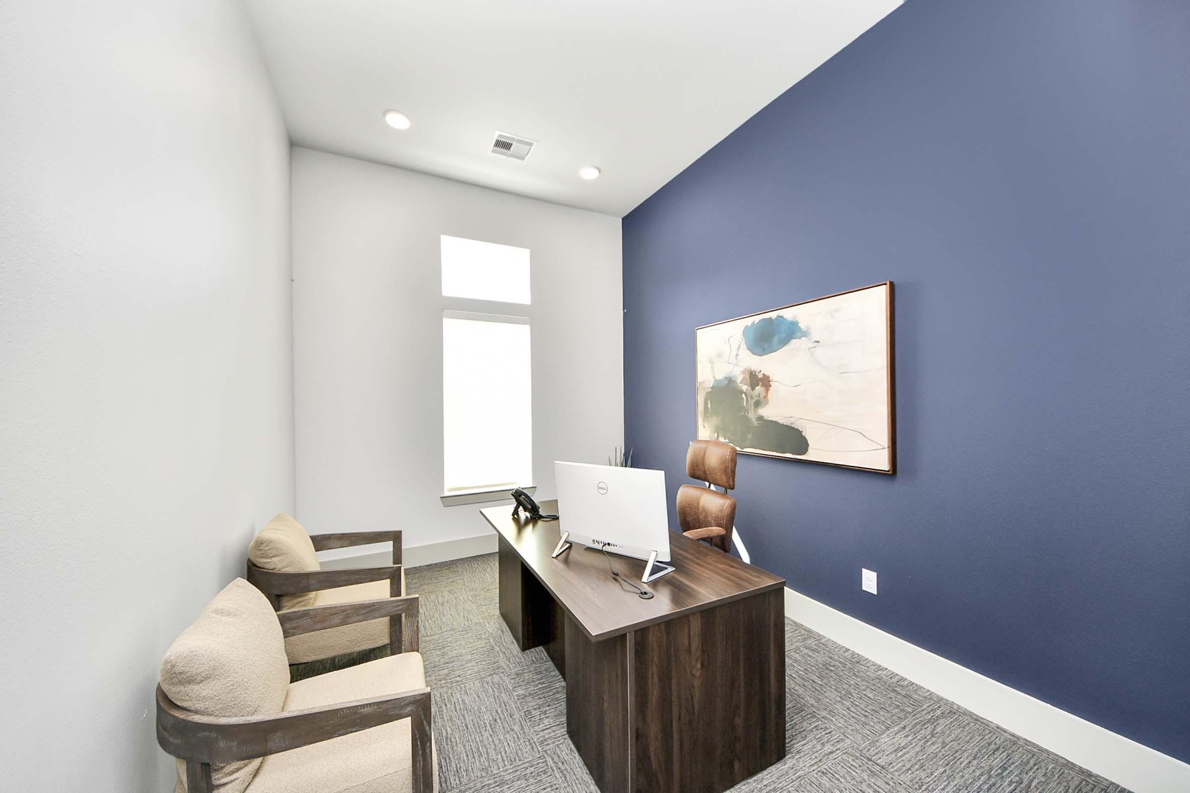 A modern office space featuring a wooden desk with a monitor, a telephone, and a decorative painting on the wall. Two beige armchairs sit beside the desk, and there is a window letting in natural light. The walls are painted in a calming dark blue, enhancing the professional atmosphere.