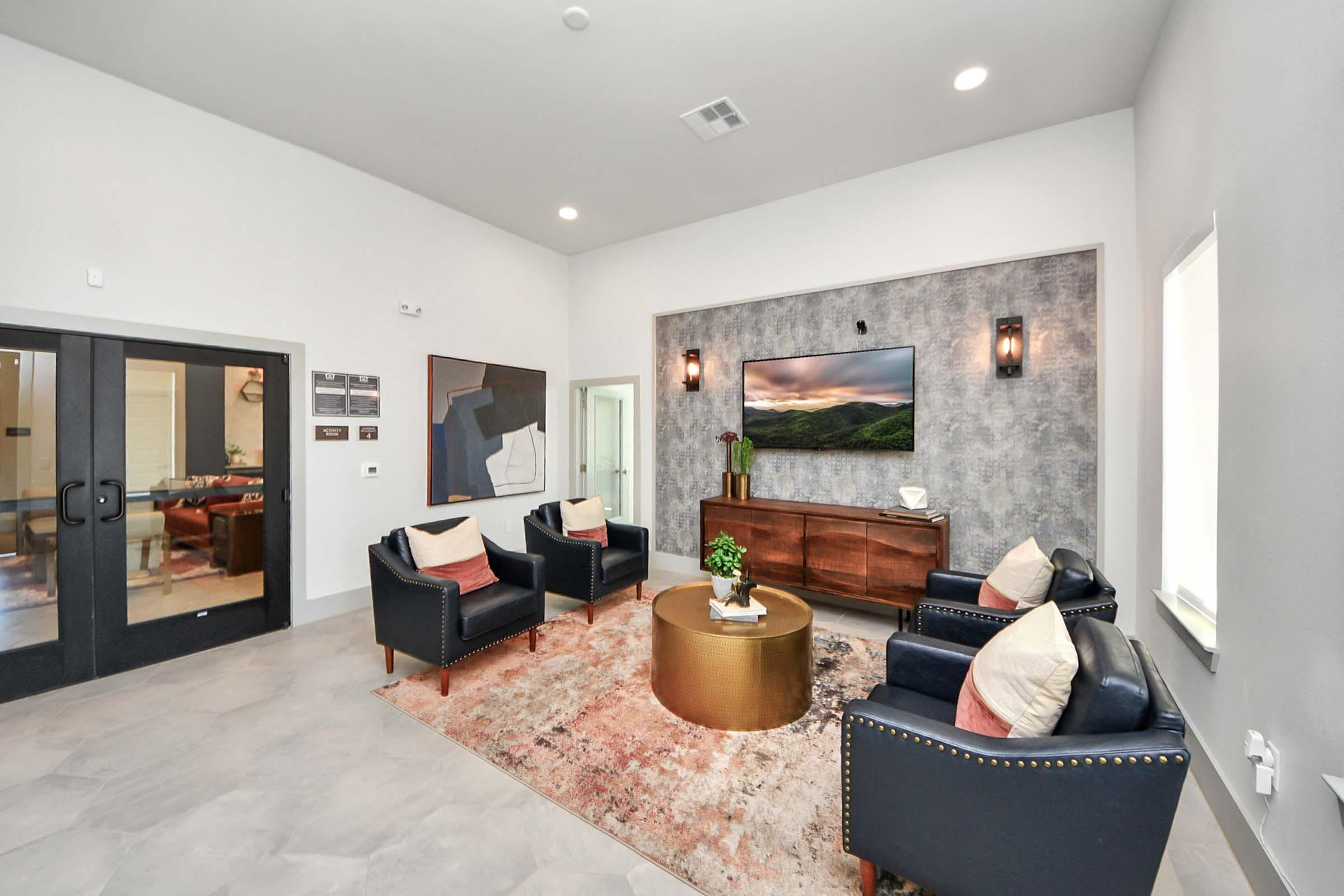 A modern living space featuring two black armchairs and a matching sofa arranged around a round coffee table. A large television is mounted on a textured wall, with a landscape image displayed. A subtle area rug adds warmth, and natural light streams through a window, while contemporary decor enhances the ambiance.