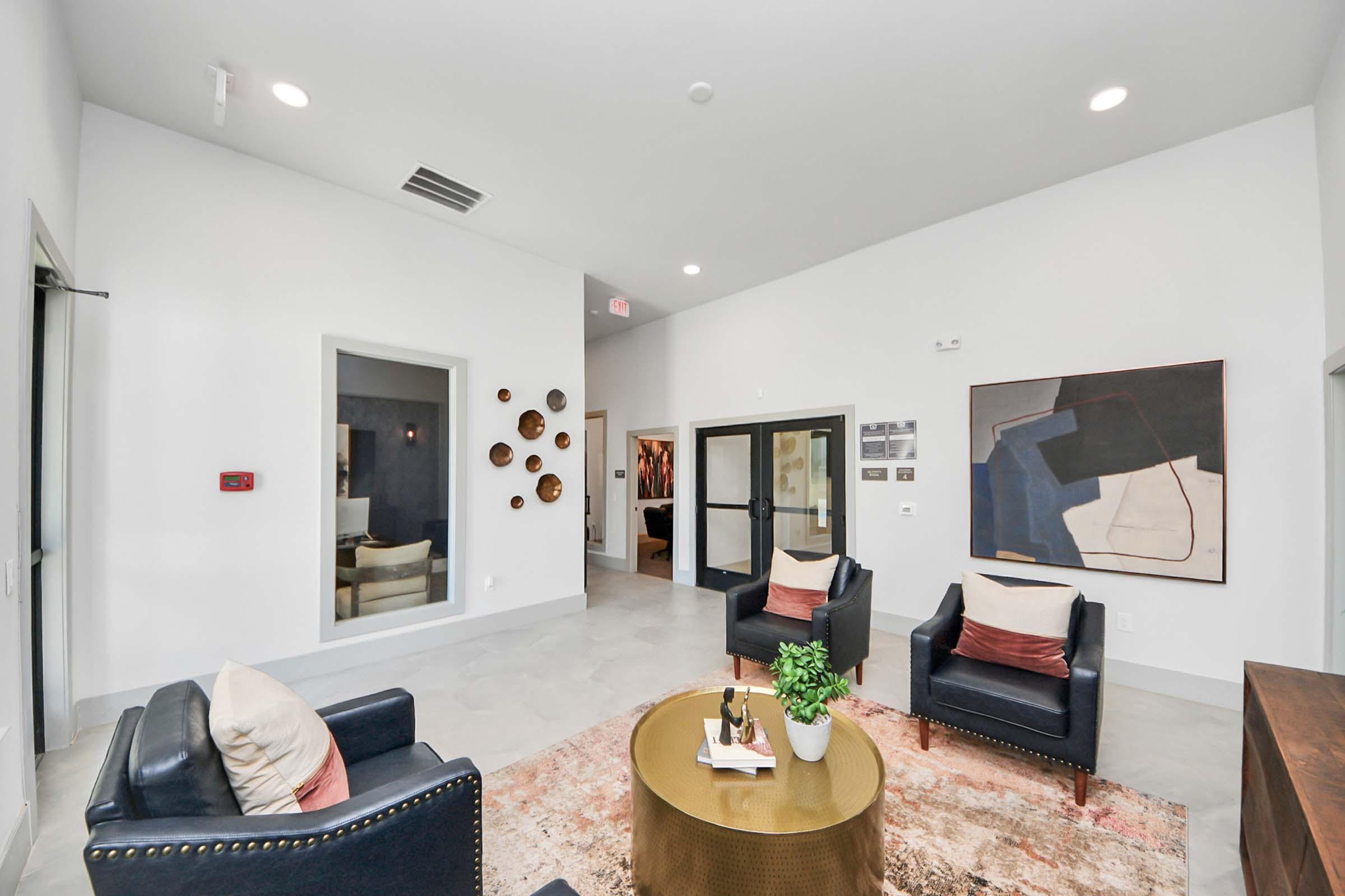 Modern interior of a spacious lounge area featuring two navy blue armchairs with pillows, a round coffee table, and a decorative rug. The walls are light-colored, with abstract artwork and wall decor. A doorway leads to another room, enhancing the inviting atmosphere of the space.