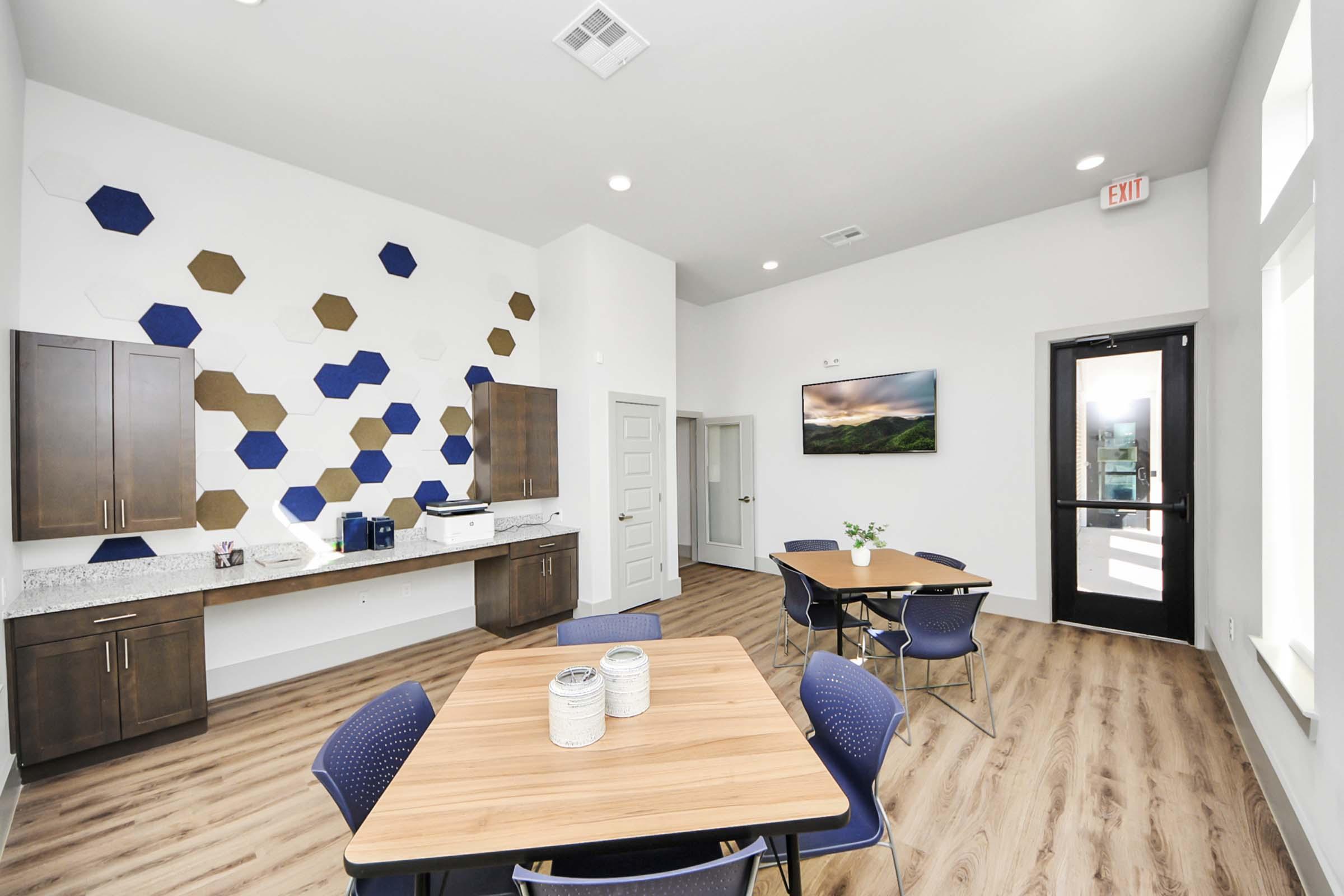 Modern interior of a communal space featuring a wooden table with blue chairs, a wall decorated with hexagonal tiles in blue and natural wood tones, a kitchenette area with dark cabinets, and a large wall-mounted screen displaying a scenic landscape. Bright lighting enhances the open feel of the room.