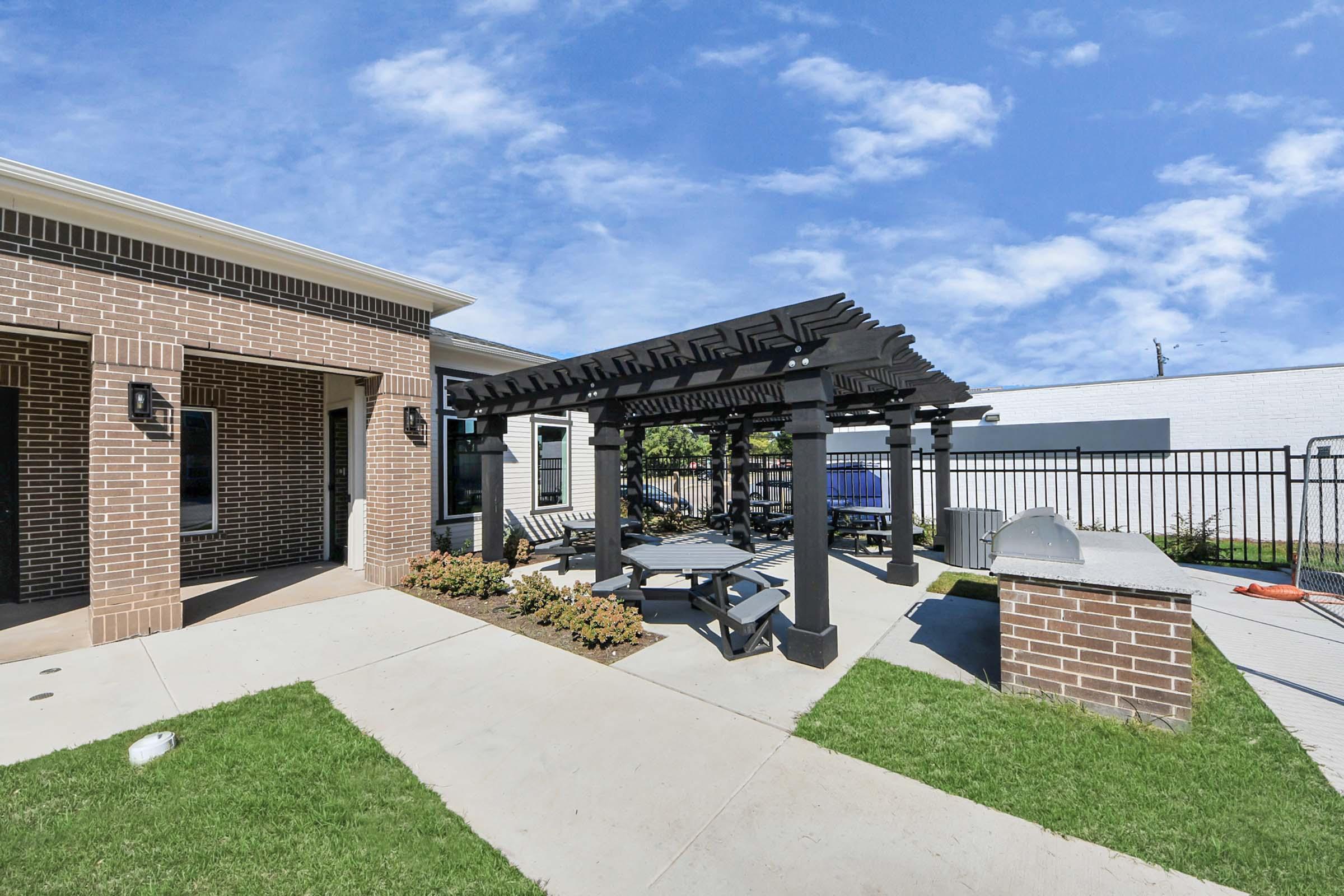 A well-designed outdoor area featuring a pergola with seating, surrounded by grass and decorative plants. The space includes a barbecue grill and two picnic tables, set against a backdrop of a residential building and a blue sky with scattered clouds.