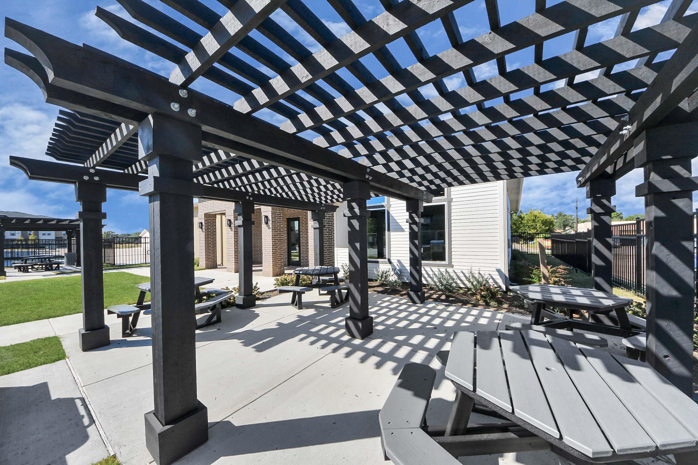 Outdoor courtyard with wooden pergolas providing shade over picnic tables. The area features a well-maintained lawn and nearby modern buildings. The sky is clear with a few clouds, creating a bright and inviting atmosphere for gatherings or relaxation.