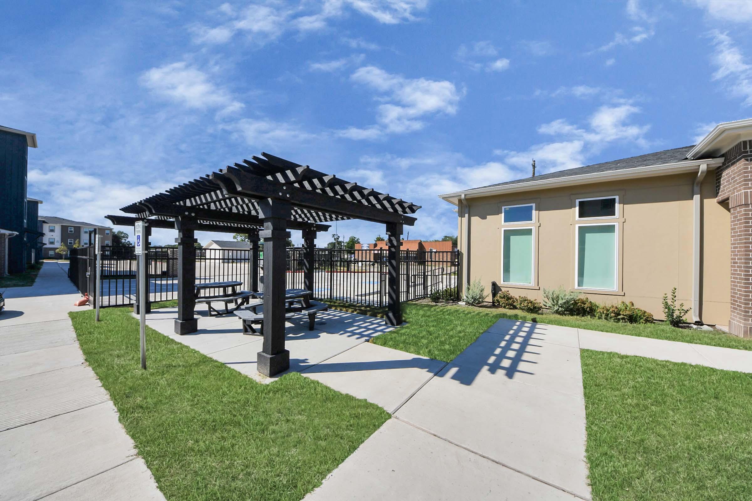 A landscaped area featuring a wooden pergola with picnic tables, surrounded by green grass. In the background, there's a modern building with large windows, under a clear blue sky with scattered clouds. The scene is well-maintained and inviting, perfect for outdoor gatherings.