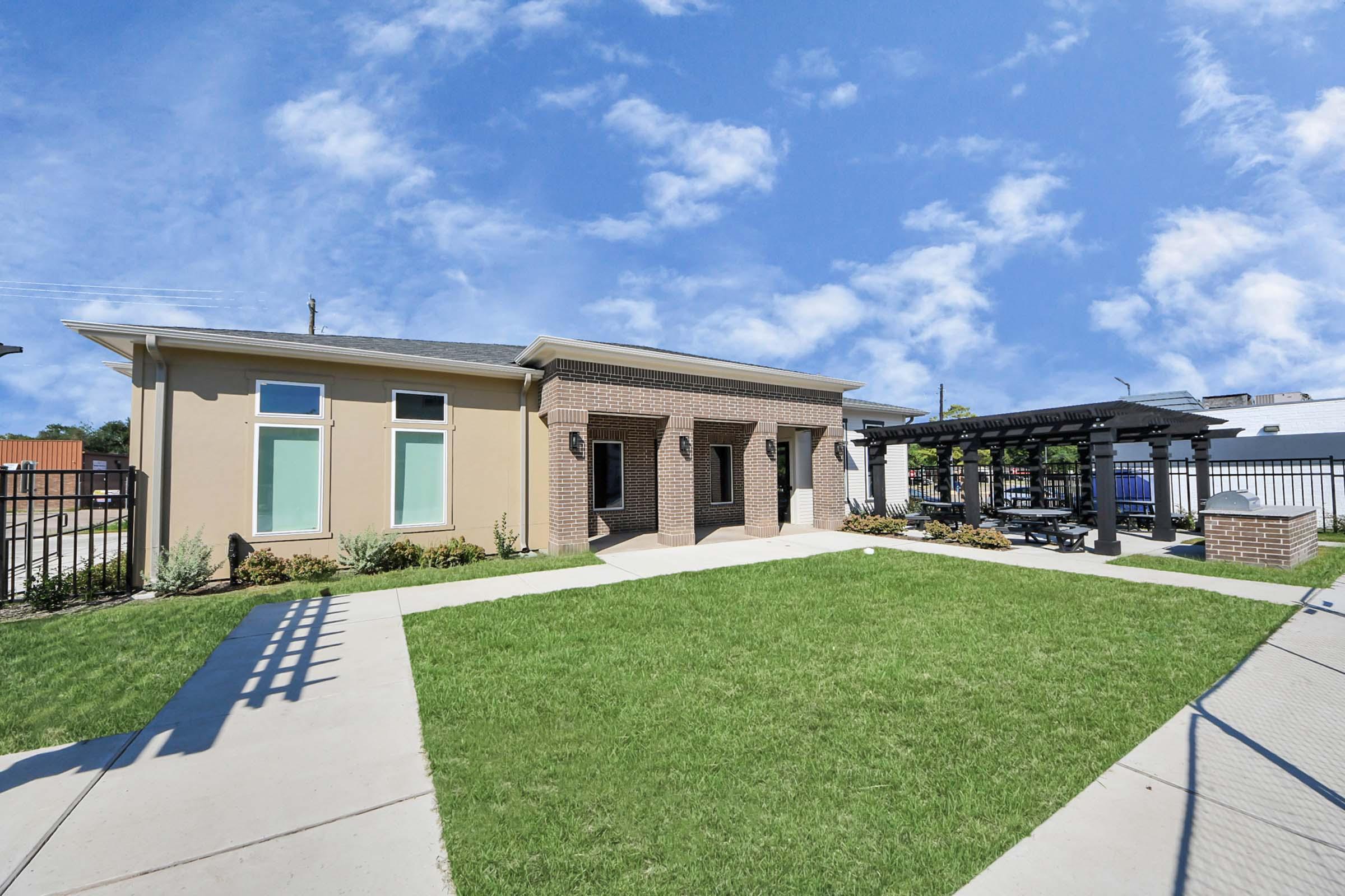 Modern building with a brick facade and large windows, surrounded by landscaped green grass. A paved pathway leads to the entrance, and there's a shaded seating area nearby with a pergola. The sky is bright blue with a few clouds, creating a welcoming atmosphere.