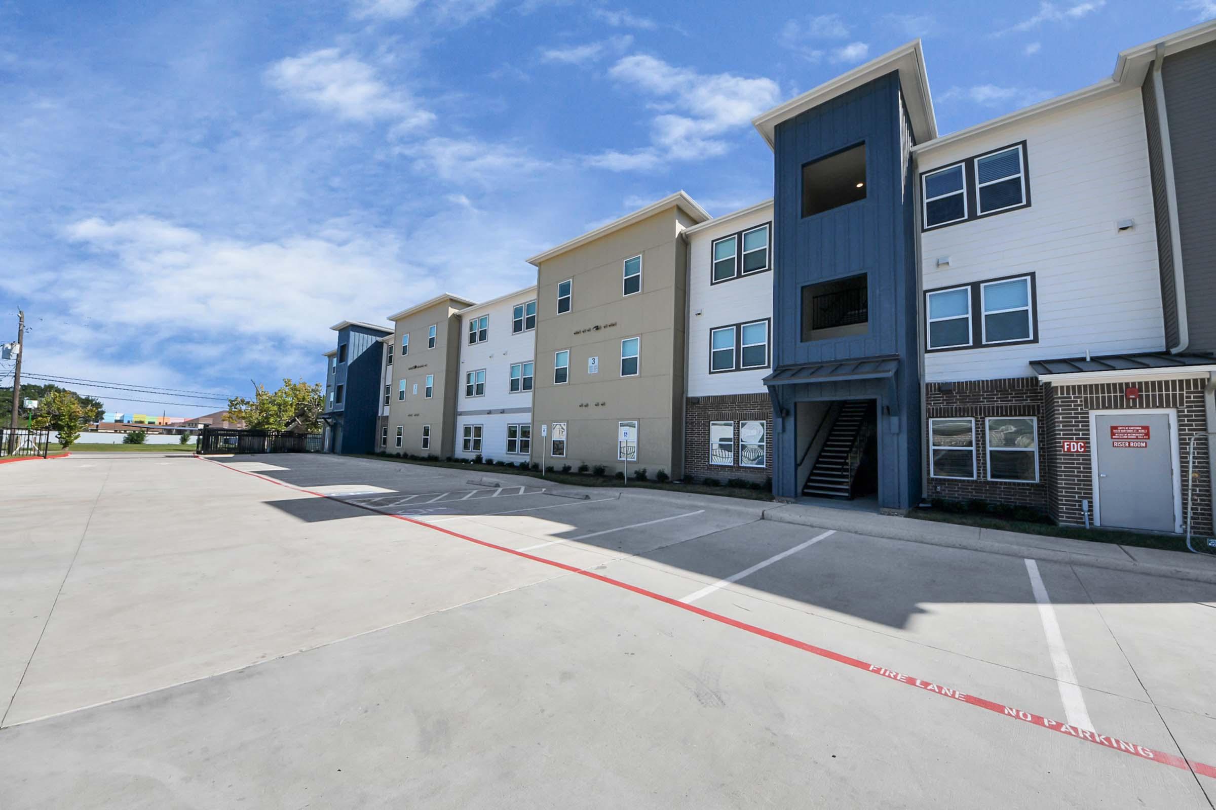 A modern multi-story apartment building with a mix of beige and blue exterior, featuring balconies and large windows. The building is situated next to a paved parking lot under a clear blue sky with a few clouds. There are marked parking spaces in front of the building.