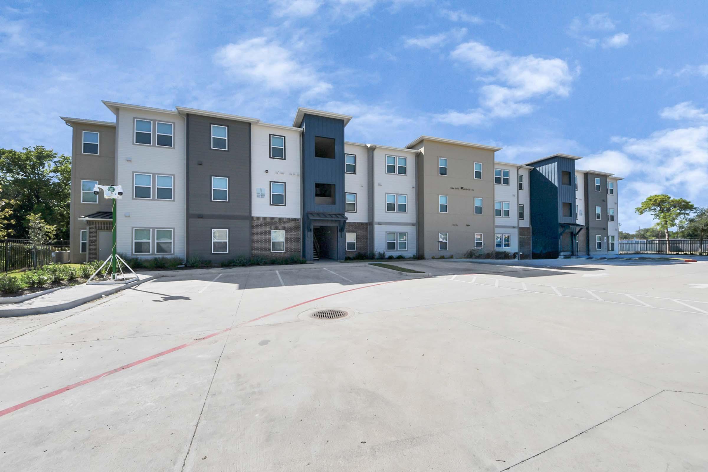 Modern multi-story apartment building with a mix of light and dark exterior colors, featuring multiple windows and a central entrance. The surrounding area is paved with a parking lot, and there are trees in the background under a clear blue sky.