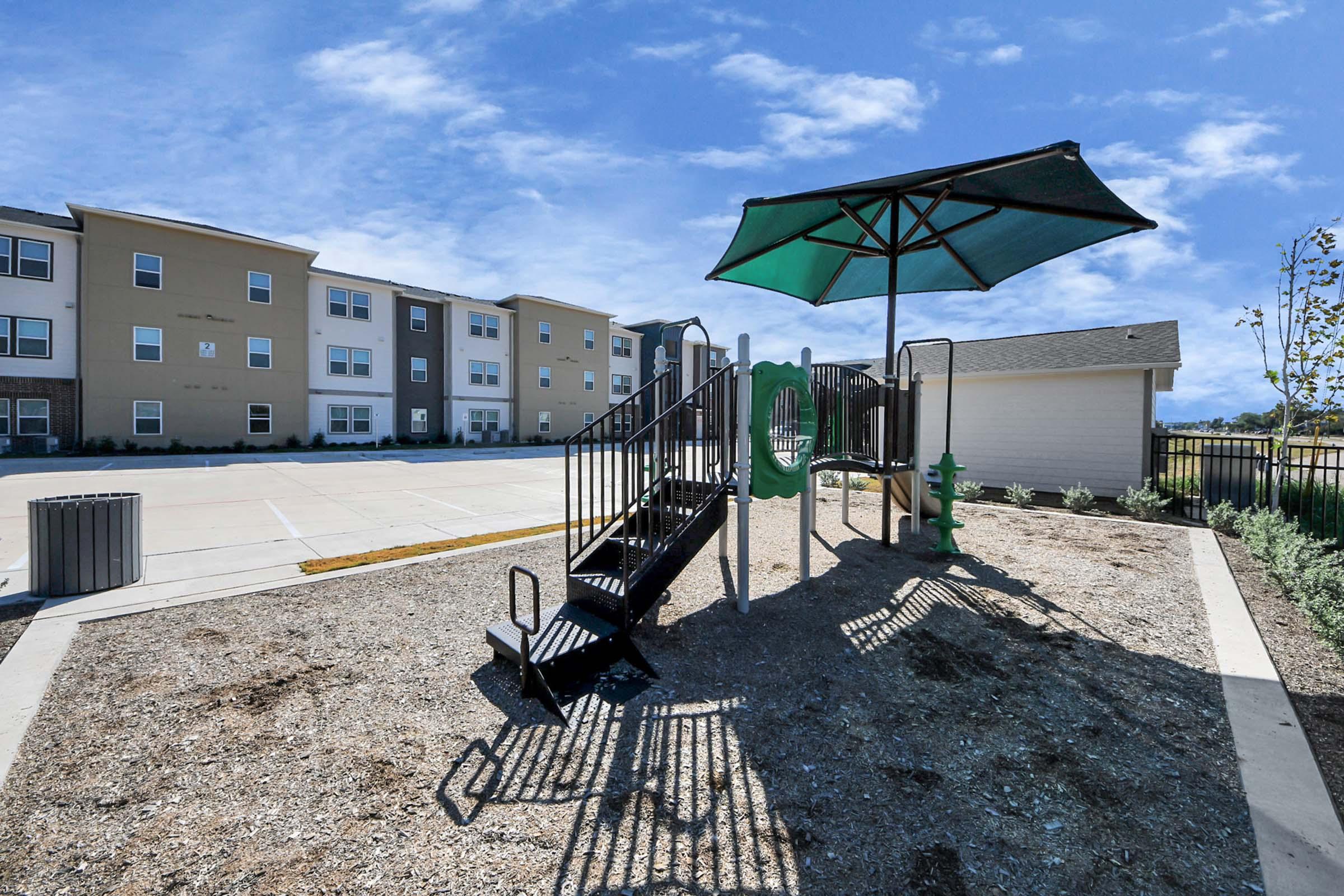 A small playground featuring a green slide and climbing structure under a large shade umbrella. The background includes several residential buildings and a clear blue sky. Gravel surrounds the playground area, and there's a trash bin nearby.