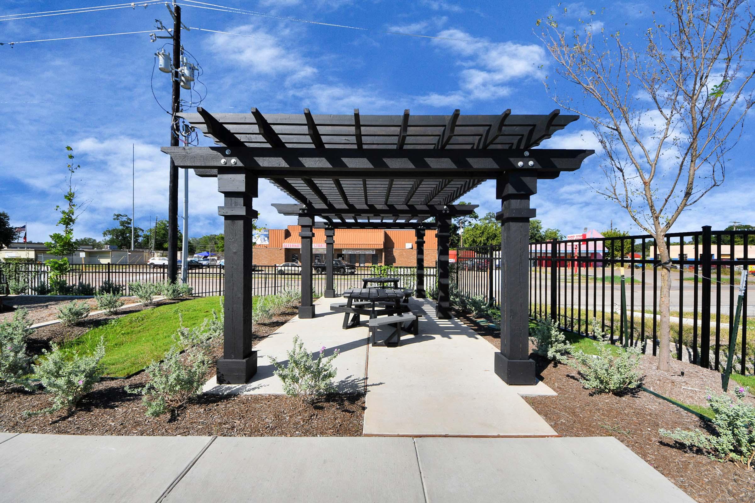 A well-designed outdoor space featuring a wooden pergola, with a picnic table beneath it. The area is landscaped with shrubs and plants, surrounded by a black fence. In the background, there are utility poles and buildings, set against a blue sky with scattered clouds.