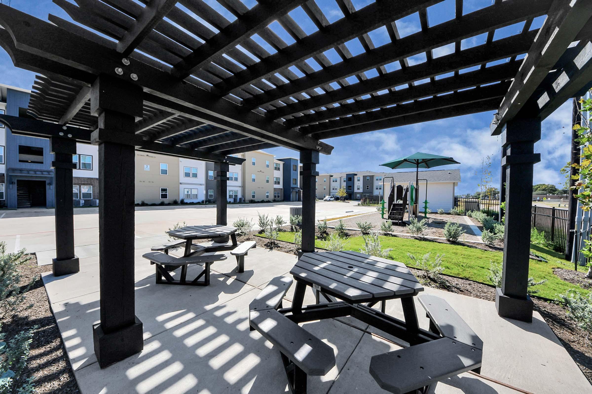 A shaded outdoor seating area featuring black picnic tables and benches under a pergola. The space is landscaped with greenery and has a playground in the background. Nearby, there is a green umbrella on the ground, providing additional shade. The setting is bright and inviting, perfect for relaxation.