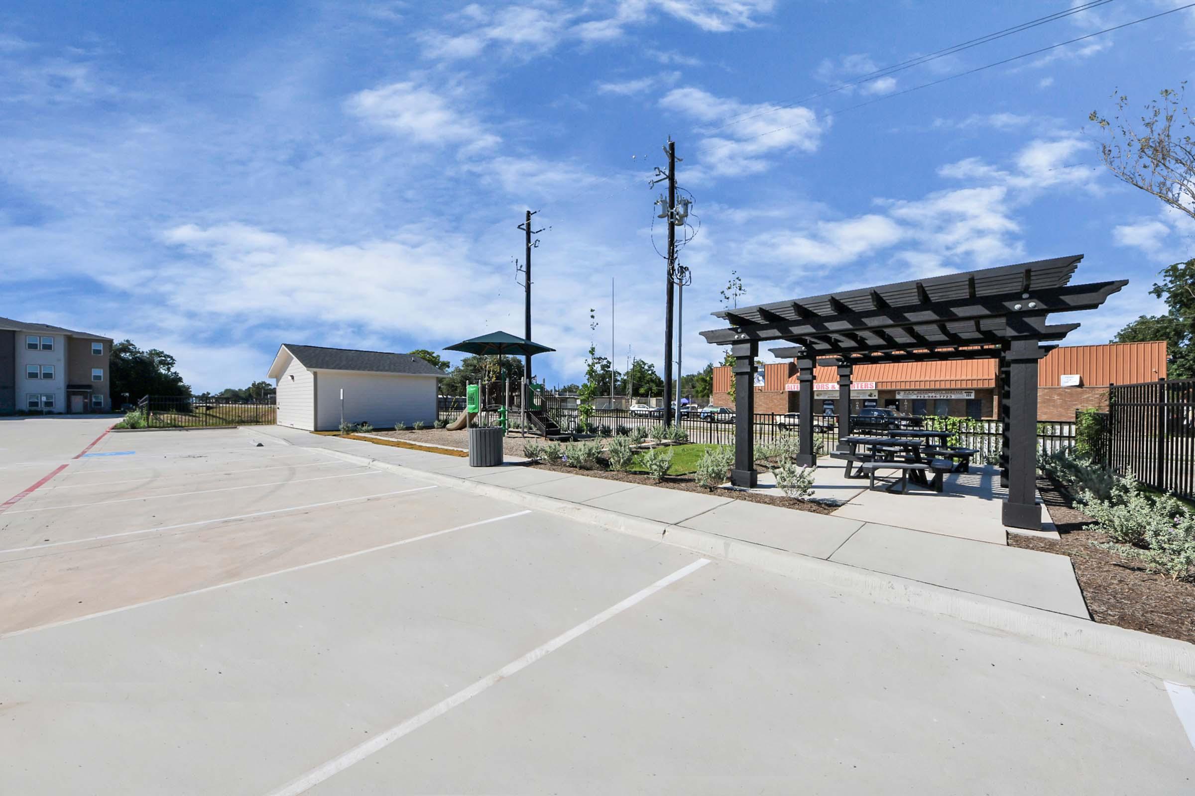 A landscaped outdoor area featuring a pergola with tables and chairs, surrounded by trees and a small grassy section. There are utility poles in the background and a designated parking area nearby, with a building visible in the distance. The sky is clear with scattered clouds.