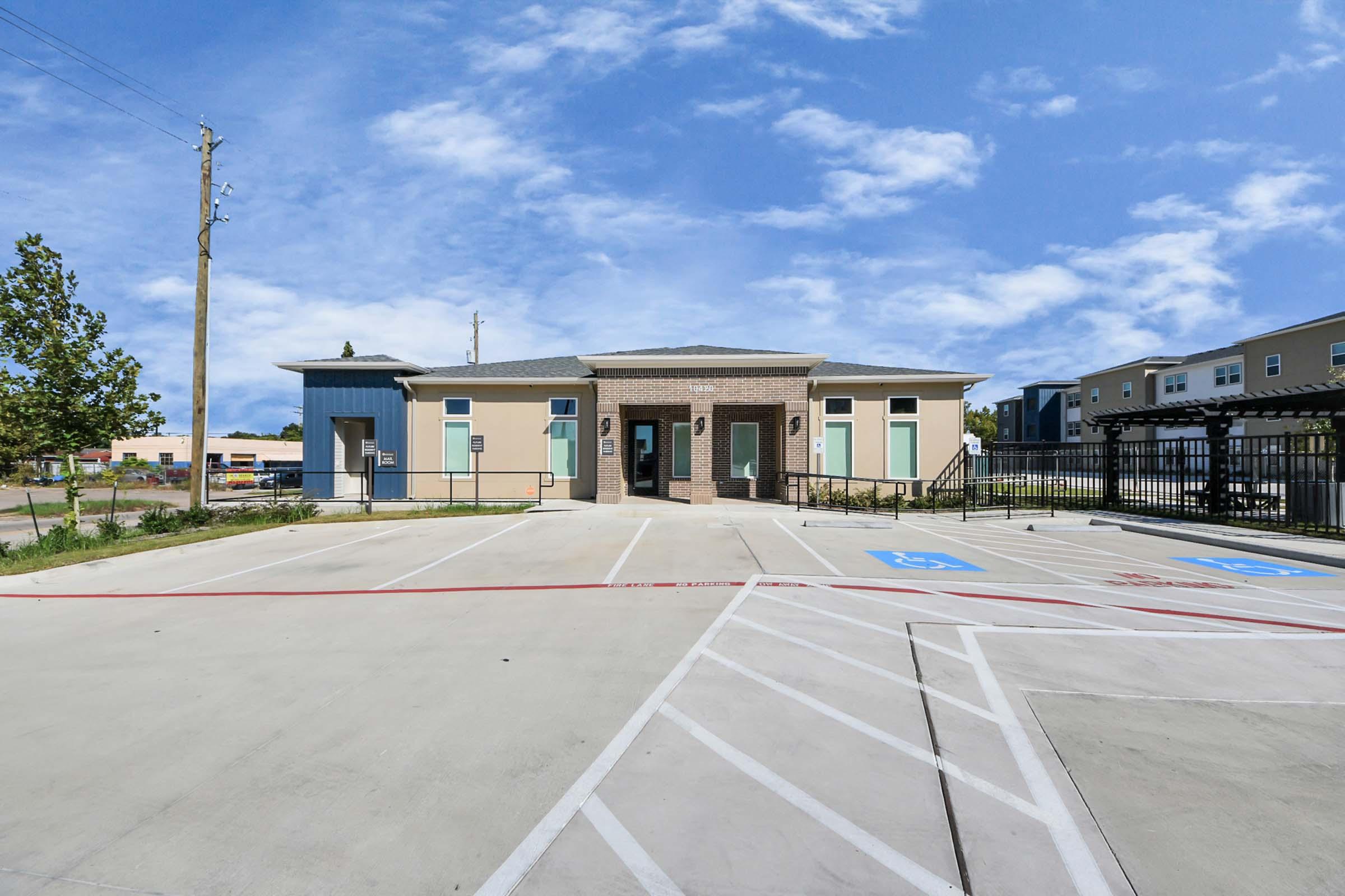 A modern, single-story building with a brick facade and light-colored siding, surrounded by a paved parking lot featuring designated parking spaces. The sky is clear with a few clouds, and there are utility poles nearby. The scene is well-lit and inviting.
