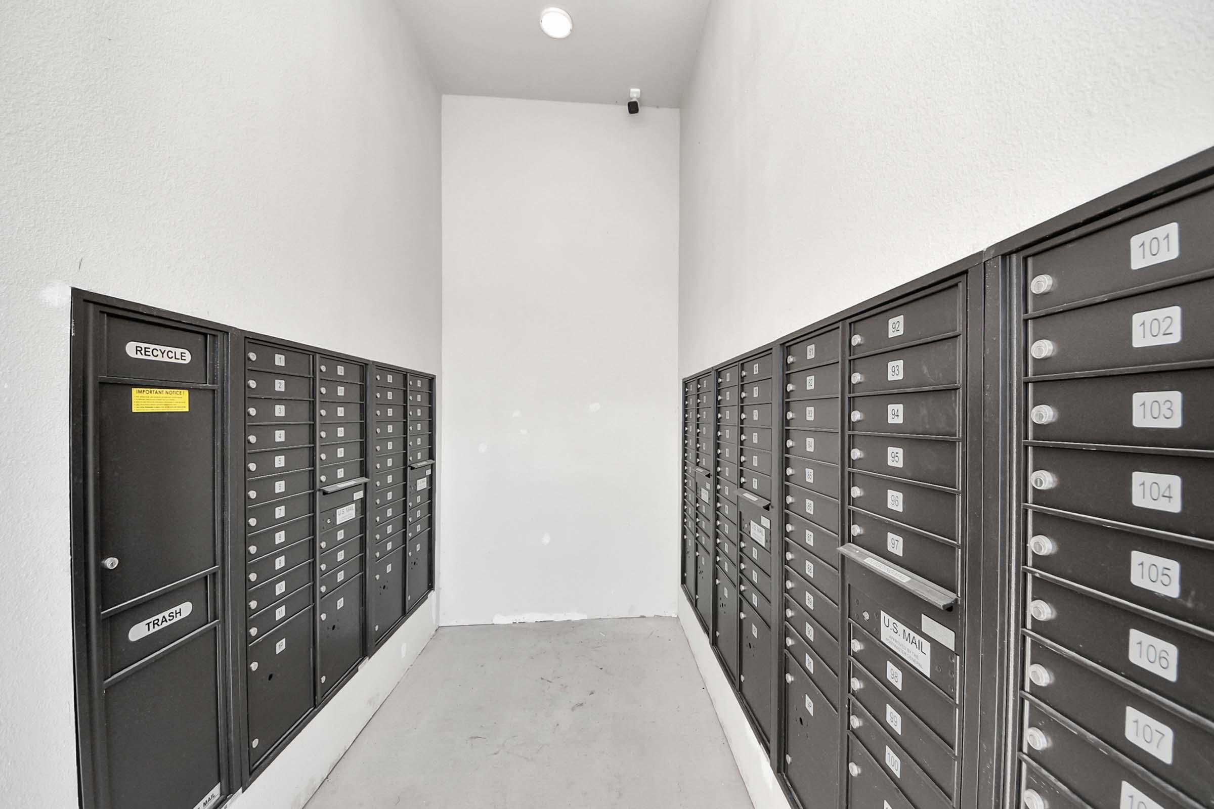 A hallway lined with black mailboxes numbered 101 to 120, with a few empty slots. The walls are white, and the space is well-lit with a ceiling light. The floor is plain and grey, and there’s a camera mounted on the wall for security.