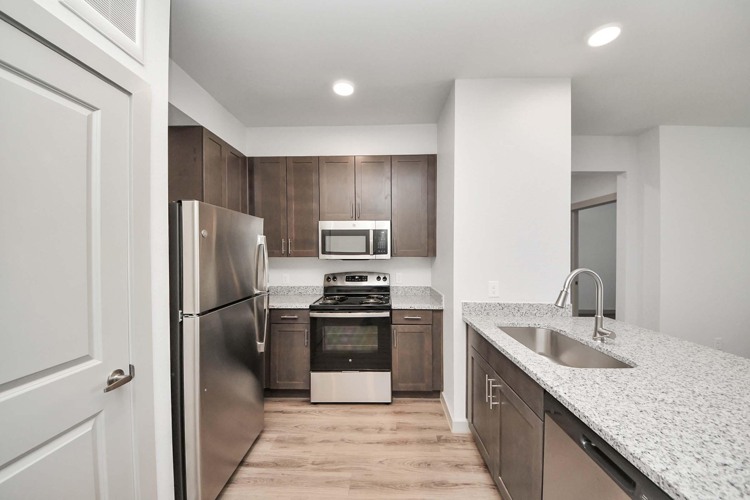 Modern kitchen featuring stainless steel appliances, including a refrigerator, microwave, and oven, set against dark wooden cabinetry. The countertop is made of light-colored granite, and the space has recessed lighting. A sink is installed in the island, with a clean, open design.