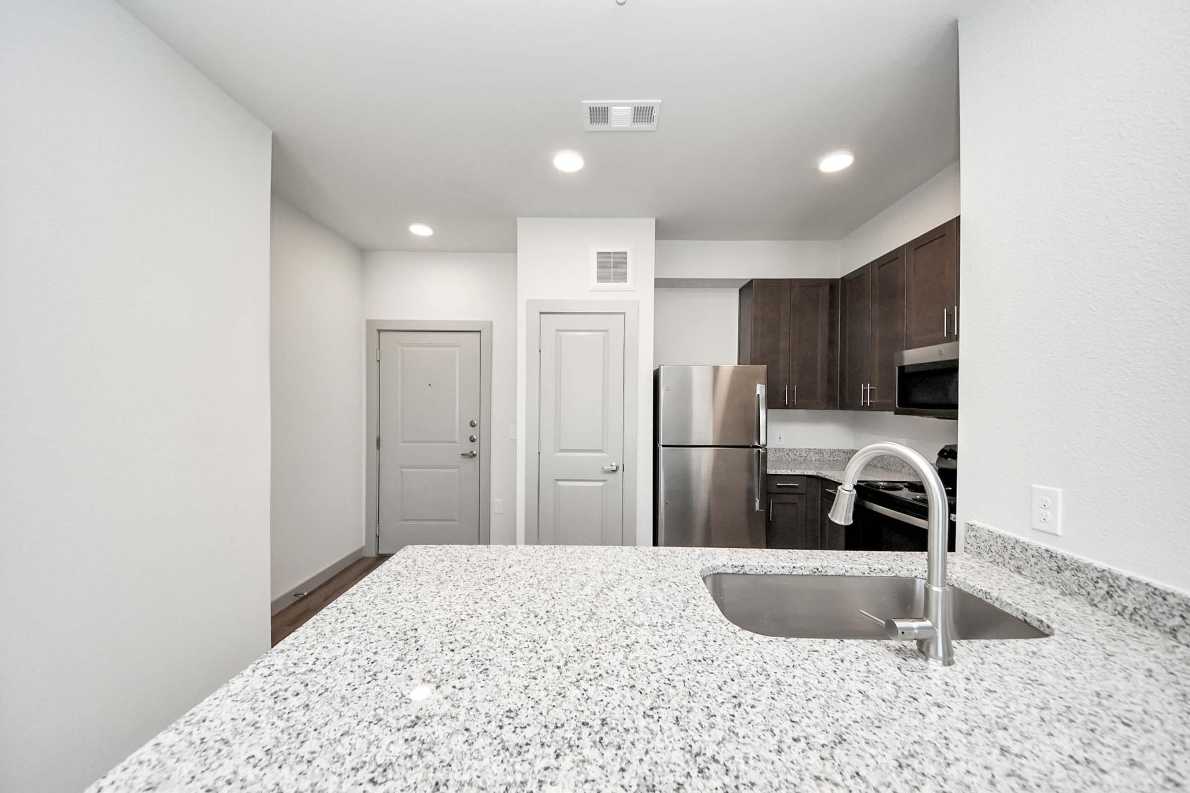 Modern kitchen featuring a stainless steel refrigerator, dark wood cabinets, and granite countertops. The space includes an undermount sink and a view into a well-lit living area with a front door visible. Neutral wall colors create a bright and inviting atmosphere.
