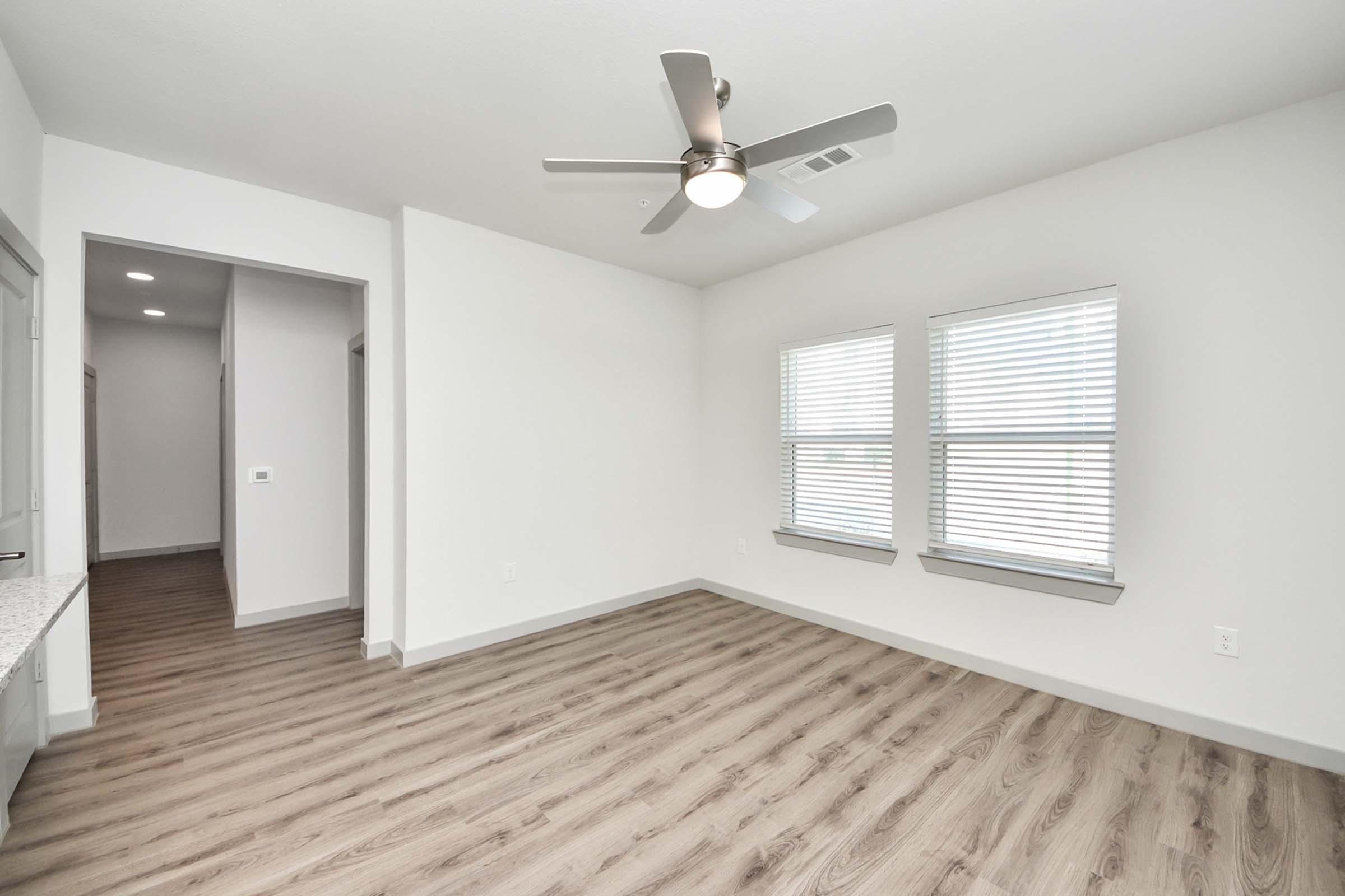 A bright, empty room with light-colored walls and a ceiling fan. It features two windows with blinds letting in natural light, and smooth, light wood flooring. A doorway on the left leads to another room, enhancing the open feel of the space.