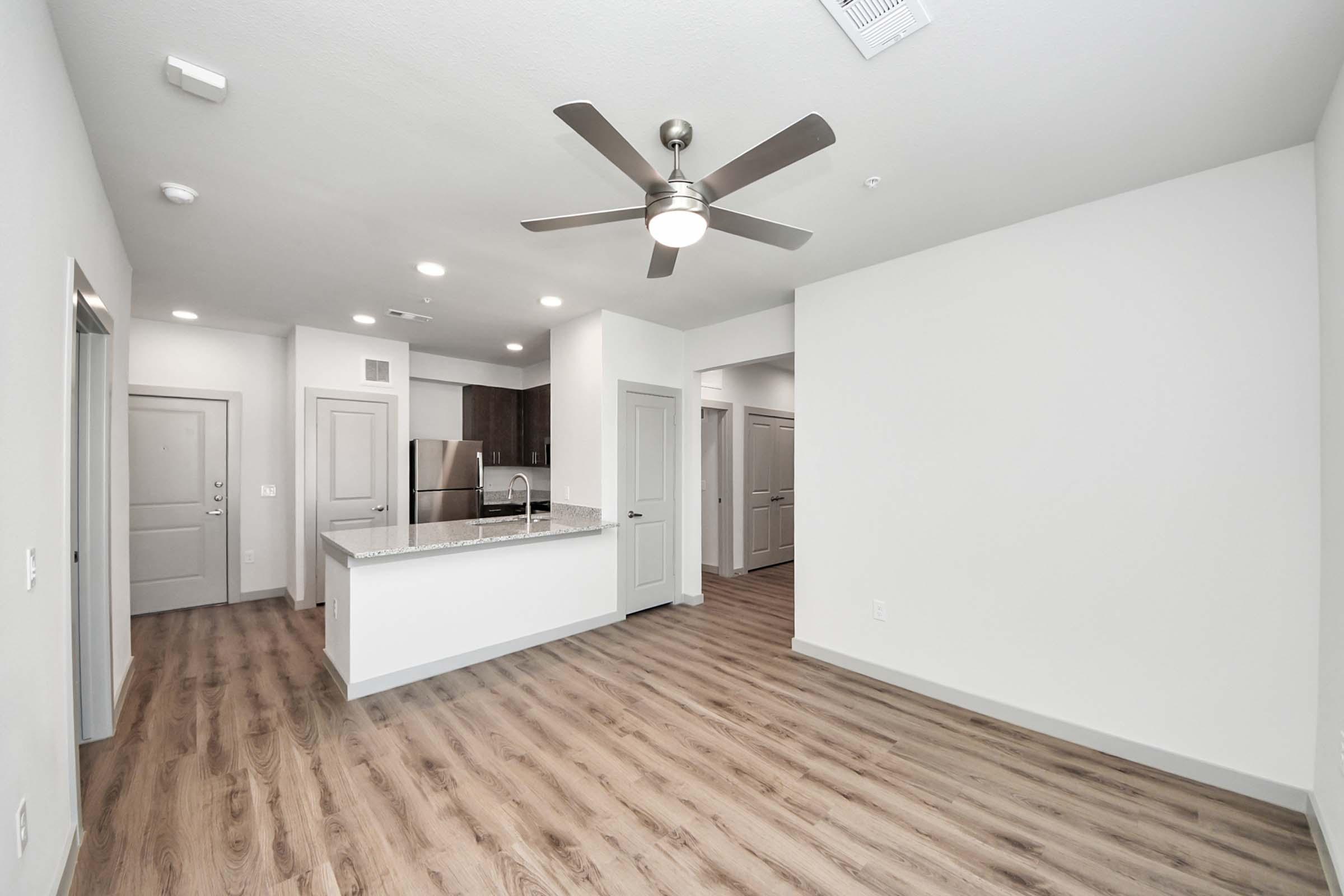 A modern apartment interior featuring an open floor plan. The living area has light-colored walls and wooden flooring. A ceiling fan is installed. The kitchen, visible in the background, has dark cabinets and a granite countertop. Doors lead to other rooms, and there is natural light from windows.