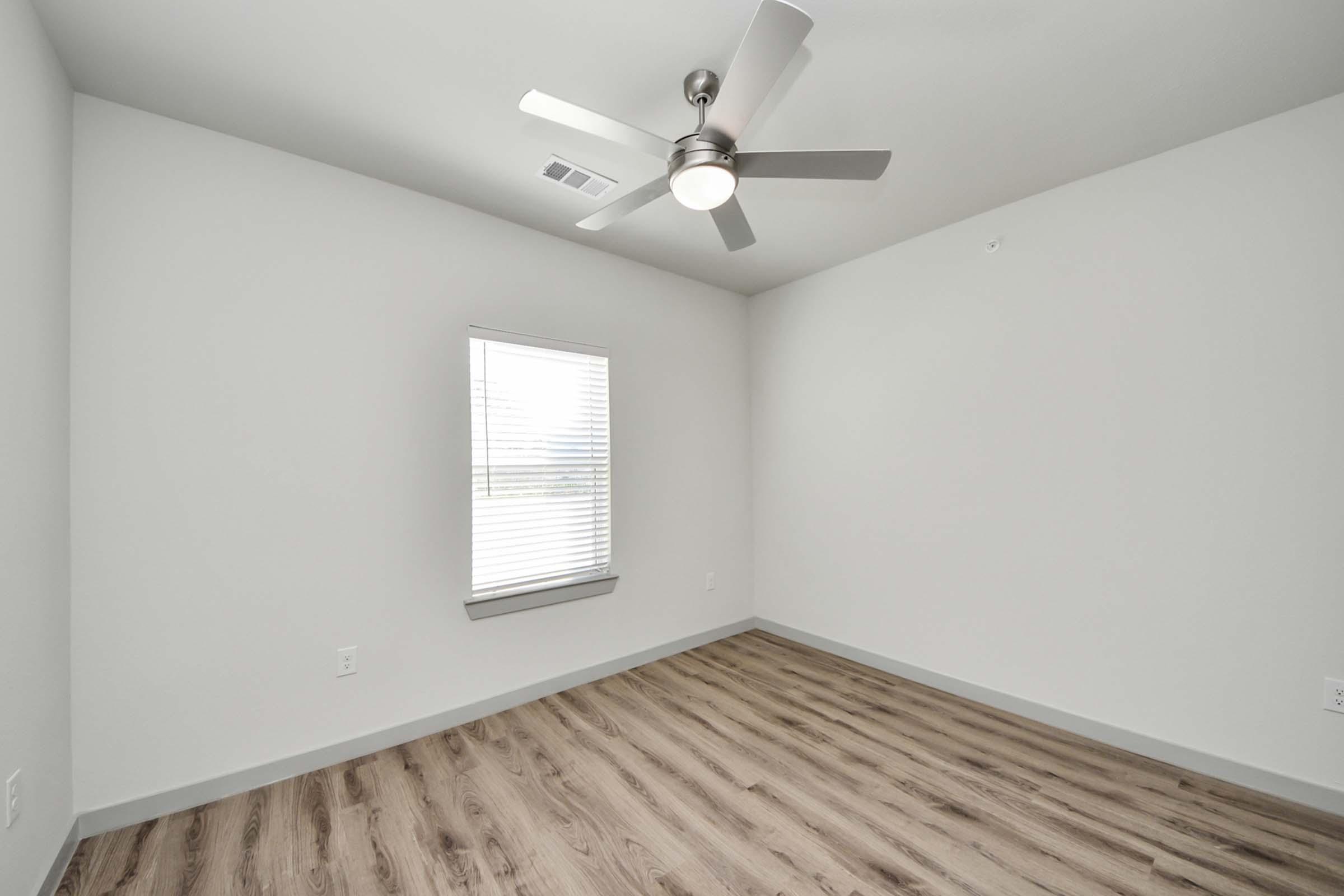 A minimalist room featuring light gray walls, a window with blinds, and a ceiling fan. The floor is covered in light wood-like laminate. The room is empty, creating a spacious and clean aesthetic, suitable for various furnishings.