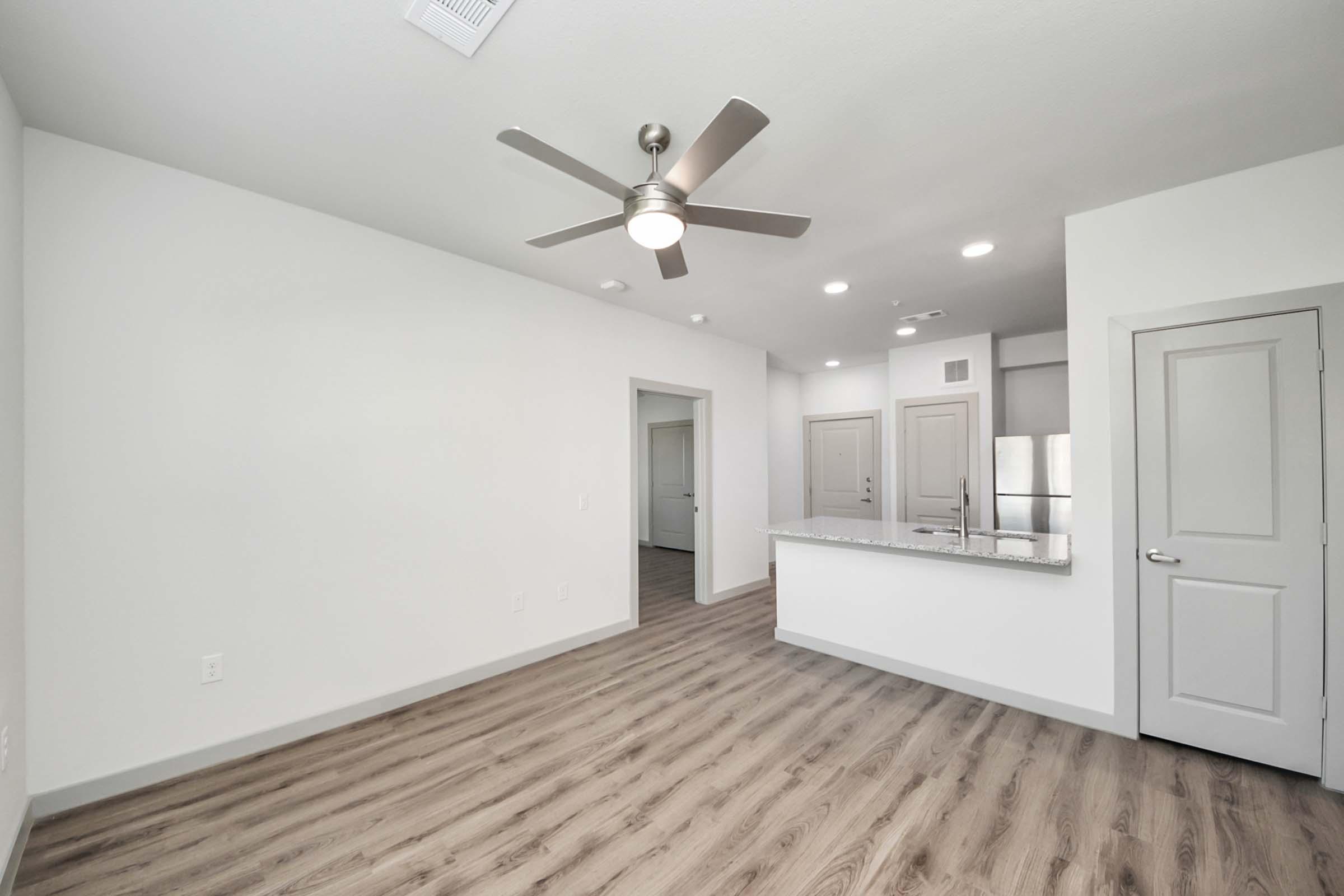 A modern, open-concept living space featuring light-colored walls, wood-like flooring, and a ceiling fan. To the left, there's an entrance to a room, and in the background, a kitchen area with a granite countertop and stainless steel appliances is visible.