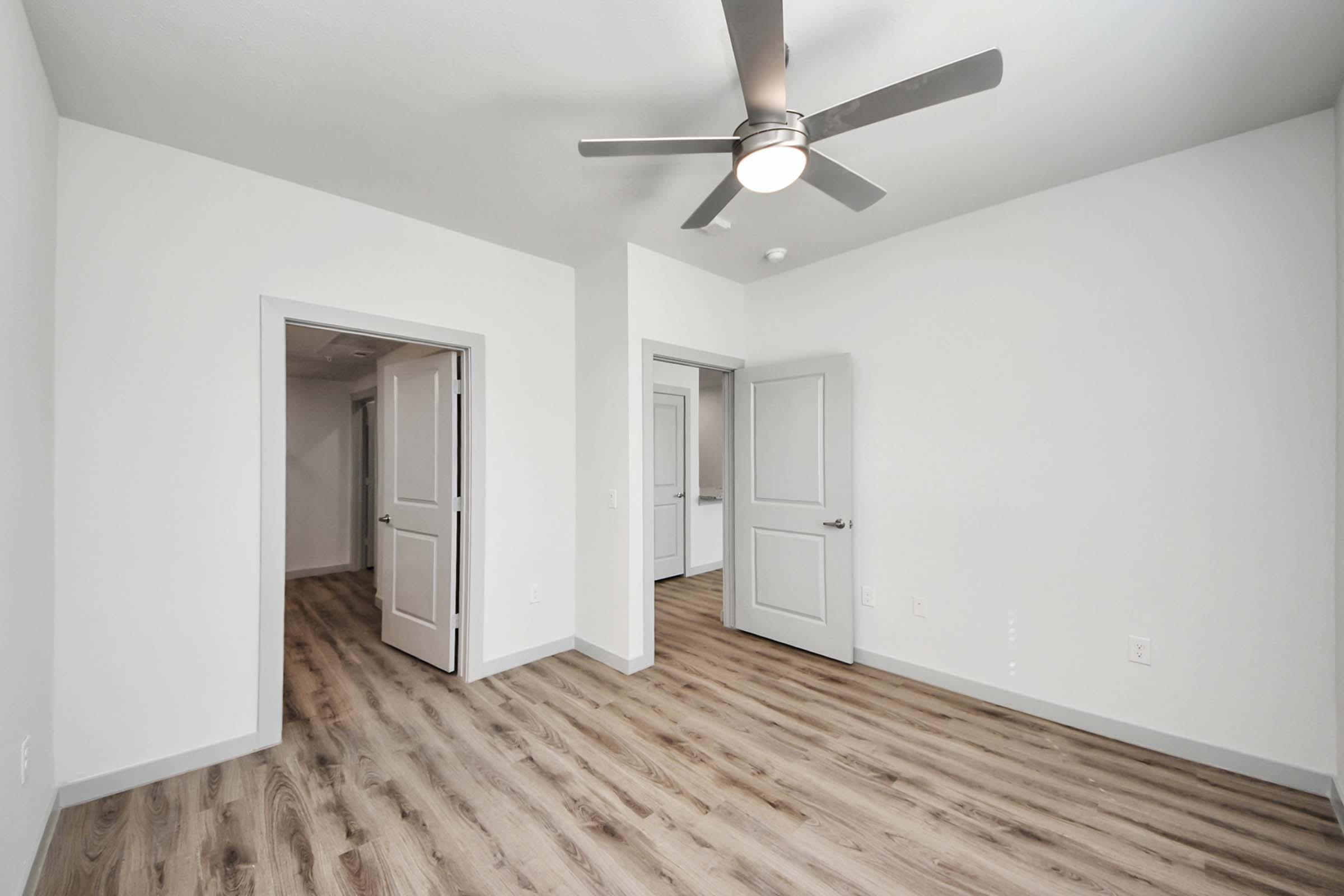 A spacious, empty room with light-colored walls and wooden plank flooring. The room features a ceiling fan and has two doorways leading to other areas. Natural light brightens the space, giving it a fresh and modern appearance.