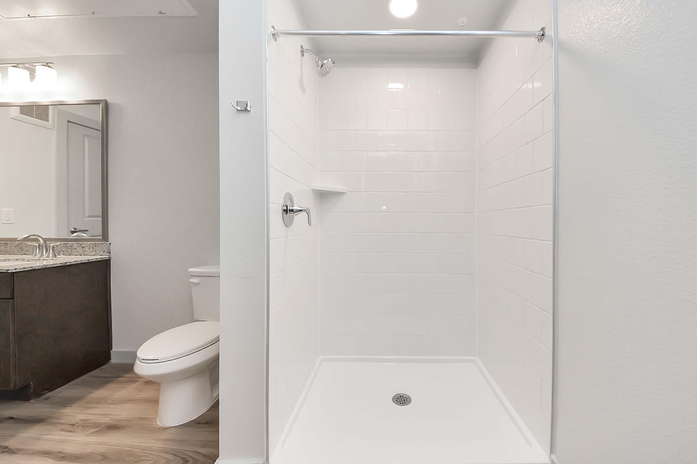 A clean, modern bathroom featuring a walk-in shower with white tiled walls, a showerhead, and a minimalistic design. To the side, there is a toilet and a countertop with a sink. The flooring is a light wood pattern, adding warmth to the space. Bright lighting enhances the overall spacious feel.
