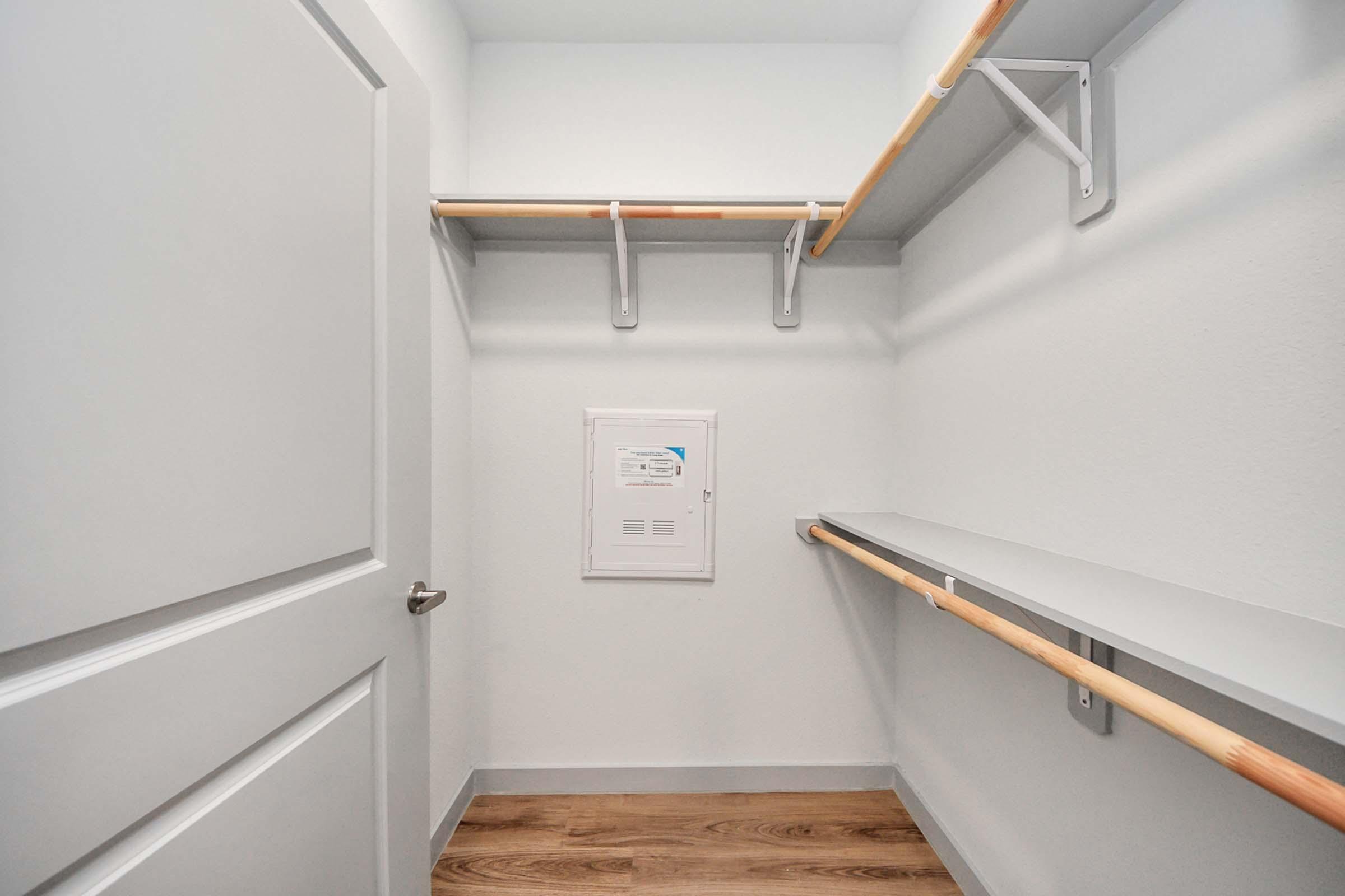 A clean, empty closet with white walls and wooden floors. It features two hanging rods for clothes, a small electrical panel mounted on the wall, and a closed door. The overall space is bright and uncluttered, suitable for storage or organization.