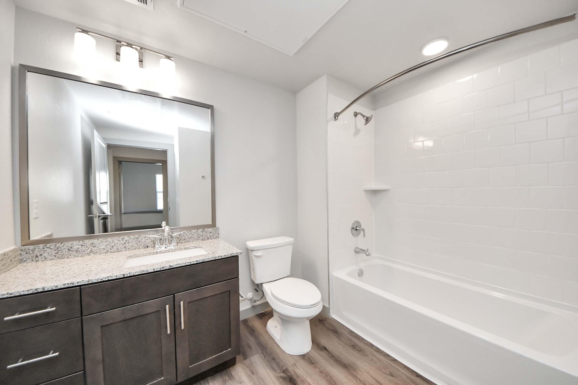 A modern bathroom featuring a white bathtub and shower, a sleek granite countertop with a sink and cabinet below, a white toilet, and a large mirror with three light fixtures above. The walls are painted in a light color, and there is a small shelf in the shower area.