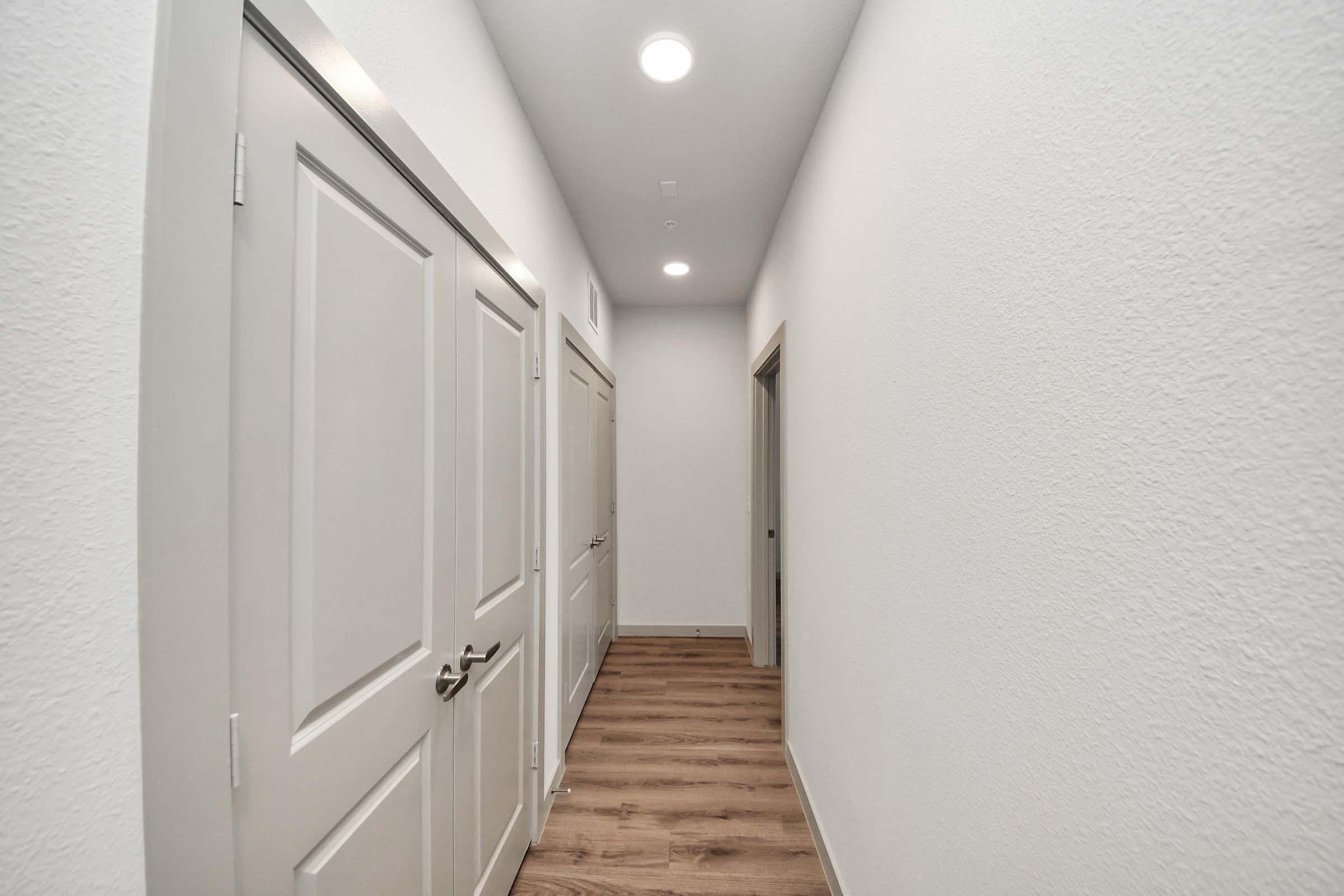 A narrow hallway with light-colored walls and wooden flooring. There are two closed doors on the left side and one door partially visible on the right. The ceiling is well-lit with recessed lights, creating a bright atmosphere in the corridor.