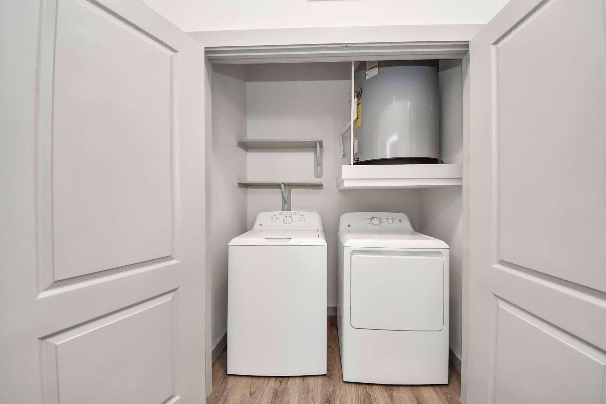 A laundry room featuring a white washer and dryer side by side. Above the washer, there is a shelf, and a water heater is visible above the dryer. The space is enclosed by double doors and has light-colored walls and wood-style flooring.