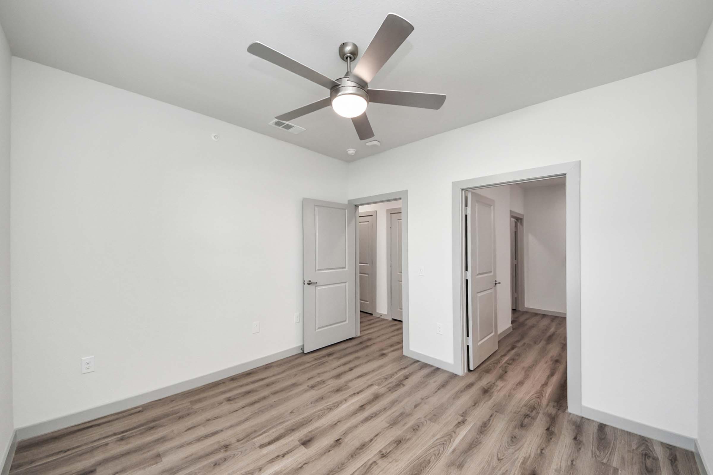 A spacious, well-lit room featuring light-colored walls and a modern ceiling fan. The flooring is a warm wood-like laminate. Two doorways lead to other areas, providing access to additional rooms. The overall design is contemporary and minimalist.