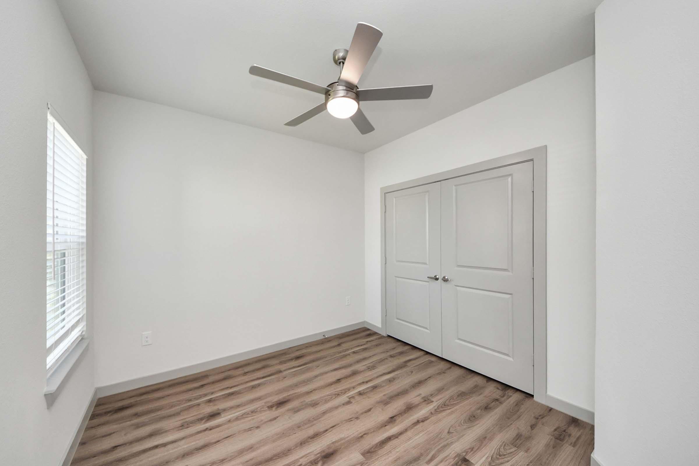 Modern interior of a bright, empty room featuring light-colored walls, a ceiling fan, and wood-like flooring. There is a window on one wall with blinds and a set of double doors opposite, providing access to storage or a closet. The space is uncluttered, emphasizing a minimalist design.