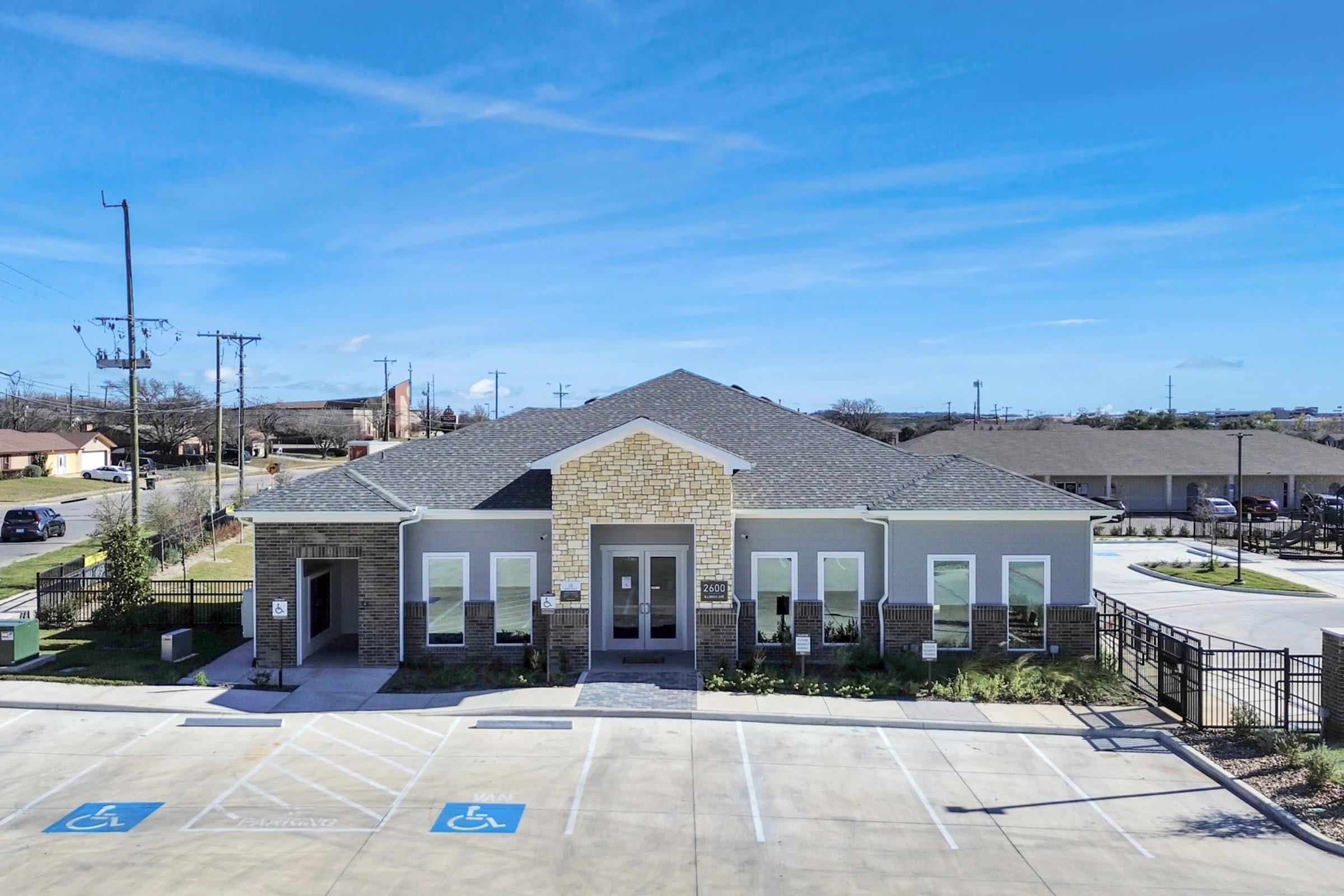 A modern, single-story building with a stone and gray exterior, featuring large windows and a pitched roof. The front has accessible parking spaces, and the surrounding area is landscaped. There are utility poles in the background and clear blue skies overhead.