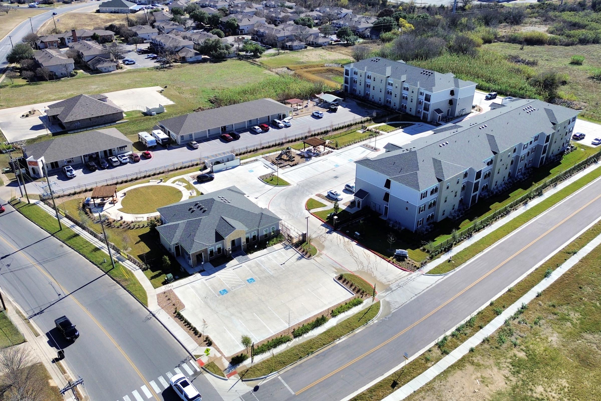 Aerial view of a residential area featuring multiple buildings. The scene shows a mixture of one to three-story apartment complexes, parking areas, and landscaped green spaces. Streets and a roundabout are visible, along with nearby undeveloped land and vegetation.