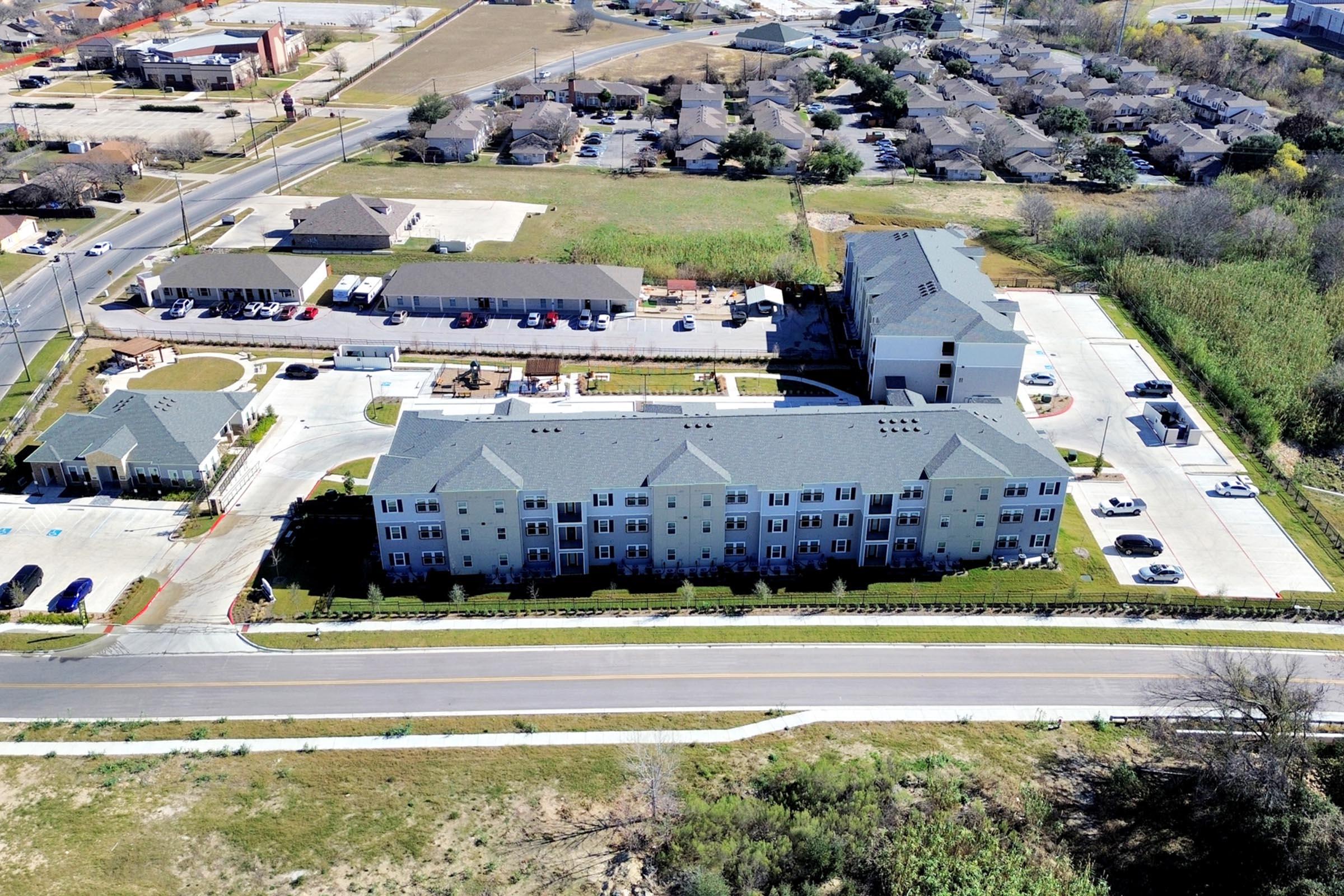 Aerial view of a multi-story residential building complex surrounded by parking lots and local roads. Nearby residential areas and a variety of facilities can be seen in the background, with green spaces and trees adding to the landscape.