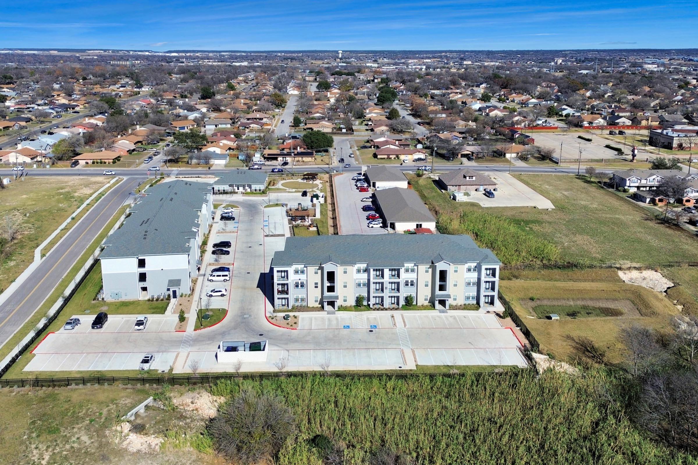 Aerial view of a residential area featuring a modern apartment complex, parking lot, and nearby buildings. In the background, single-family homes are visible, along with open fields and roads. Clear blue sky above with few scattered clouds.