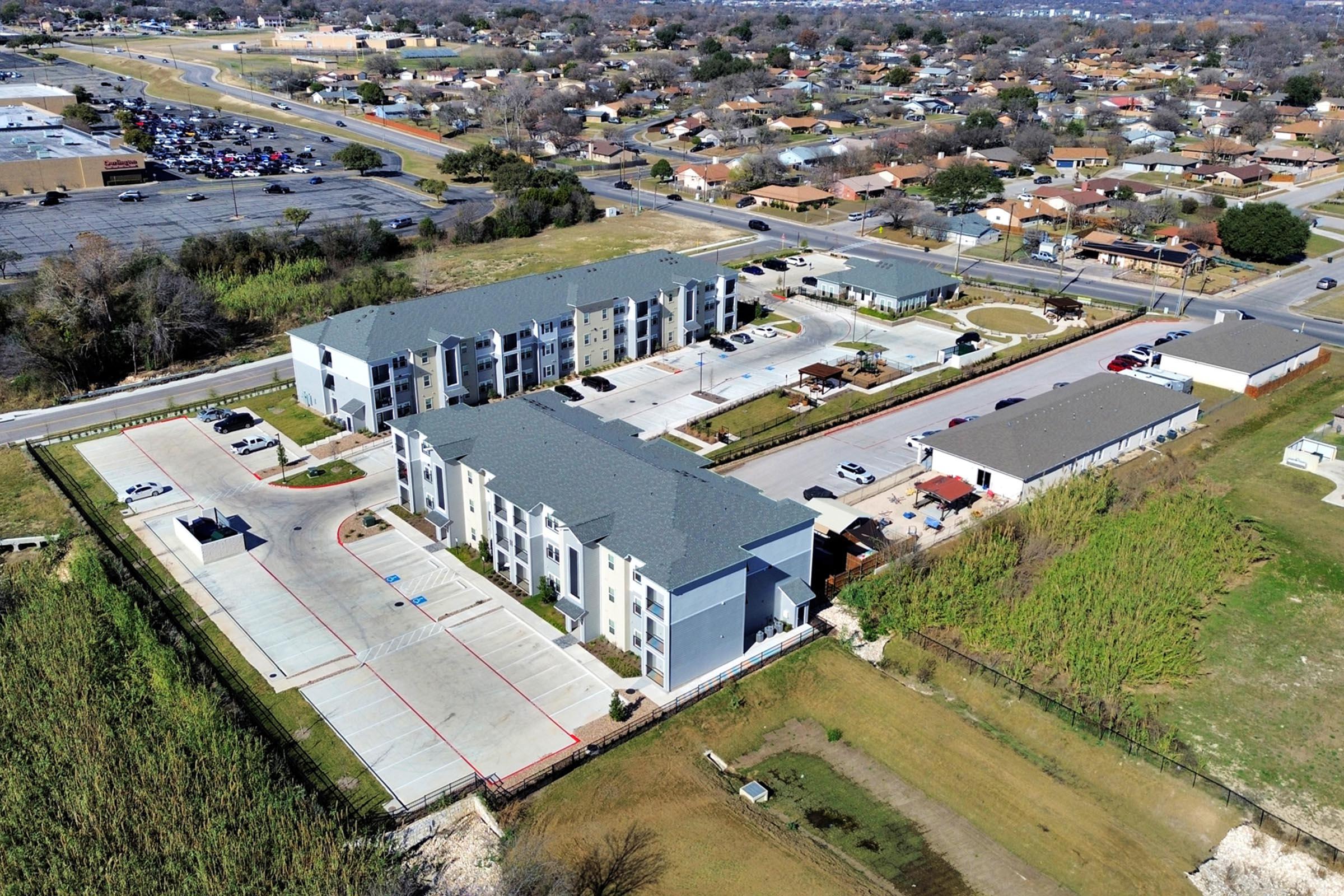 Aerial view of a residential complex featuring multiple three-story buildings with grey roofs, parking areas, and landscaped grounds. Surrounding the complex are various single-family homes and a commercial area in the distance, showcasing a suburban neighborhood.