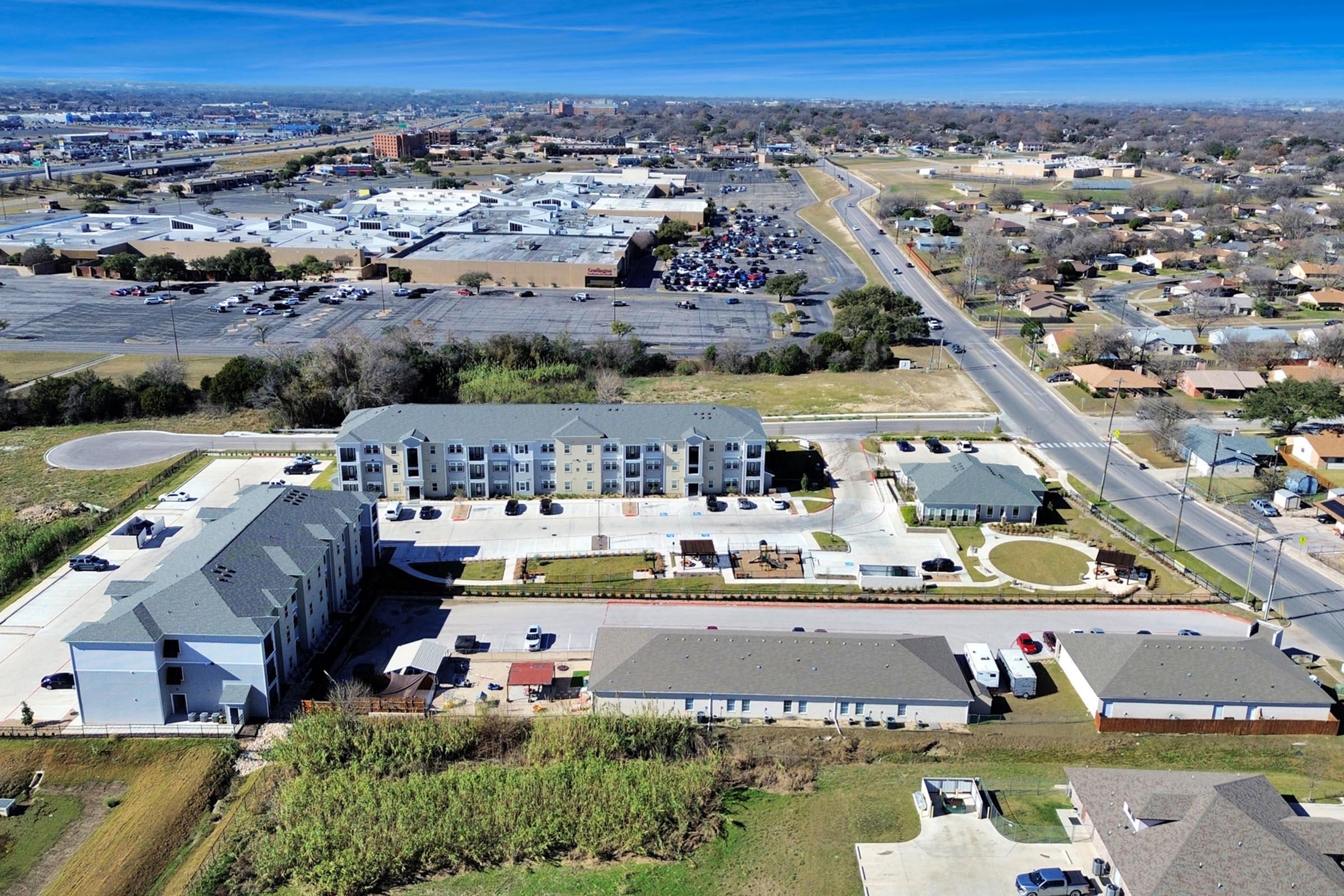 Aerial view of a residential complex with multiple buildings, surrounded by parking lots and green spaces. Nearby, a busy road runs alongside a shopping area in the background. The landscape features a mix of commercial and residential properties with clear blue skies above.
