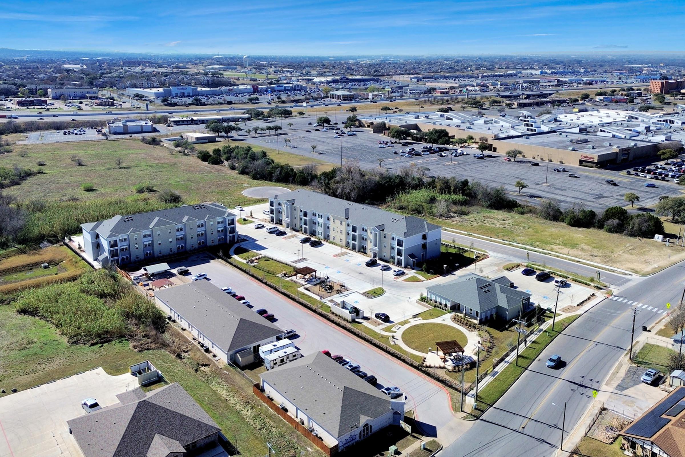Aerial view of a suburban area featuring multiple residential buildings, including an apartment complex with parking lots. Surrounding landscape includes open fields, roads, and a shopping area in the distance under a clear blue sky.