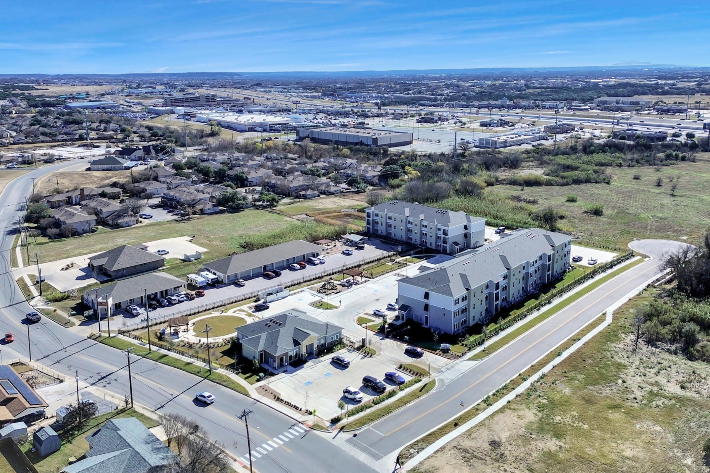 Aerial view of a suburban area featuring a new apartment complex near an intersection. Surrounding the buildings are residential homes and green spaces, with commercial facilities visible in the distance. The landscape includes roads and parking areas, under a clear blue sky.